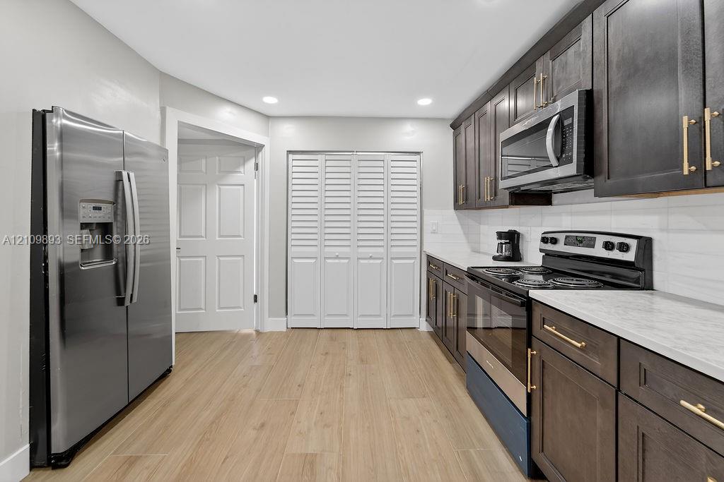 Kitchen featuring wood-finish flooring, dark-toned cabinetry with brass-finish hardware, stainless steel appliances, light-toned countertops, and a white subway tile backsplash