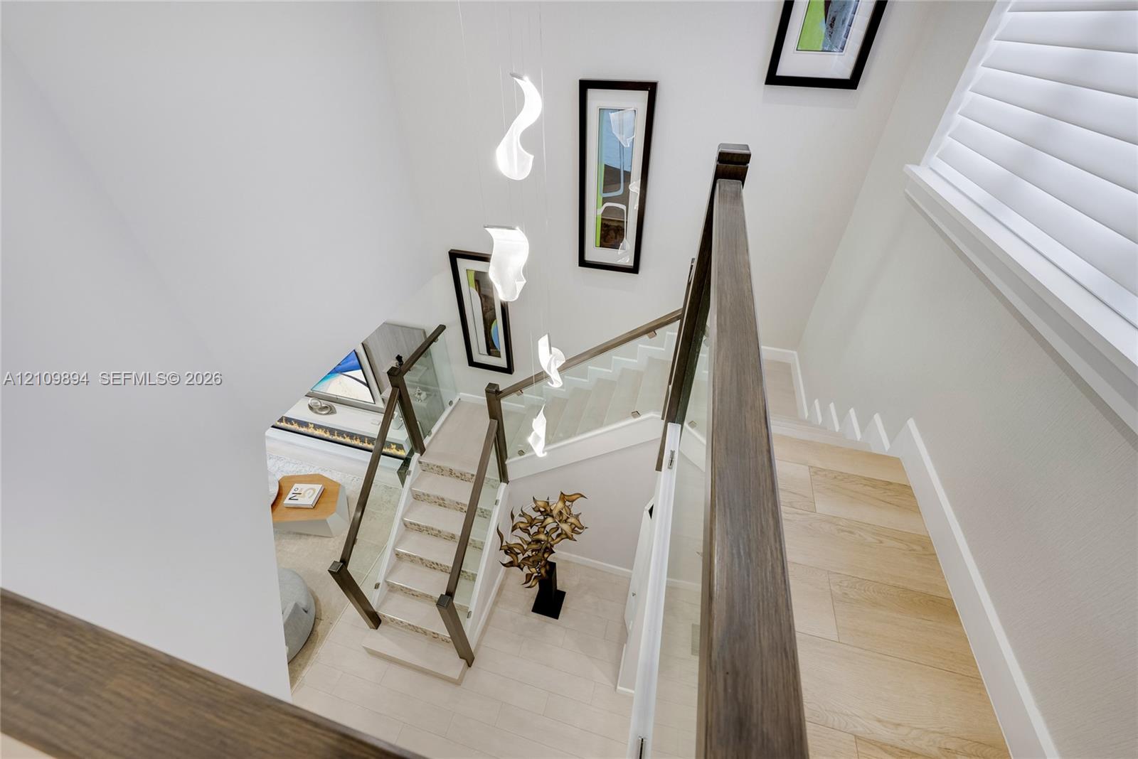 Staircase featuring wood-finish treads, glass balustrades with dark wood handrails, wood-finish flooring, white plantation shutters, and a suspended multi-light fixture