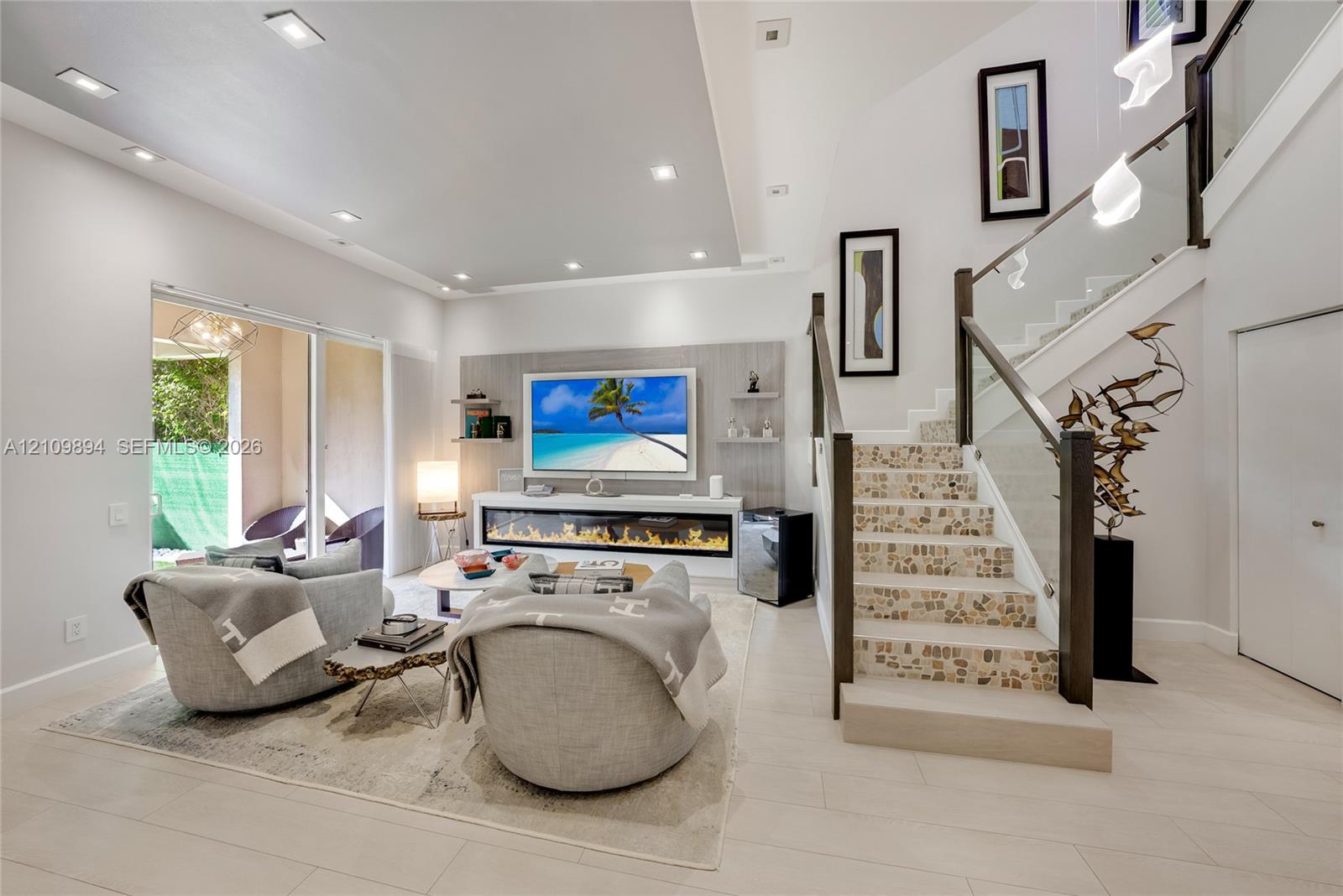 Open-concept living space featuring a recessed ceiling with integrated lighting, built-in linear fireplace with stone-finish surround, staircase with glass panel railing, and large sliding glass door