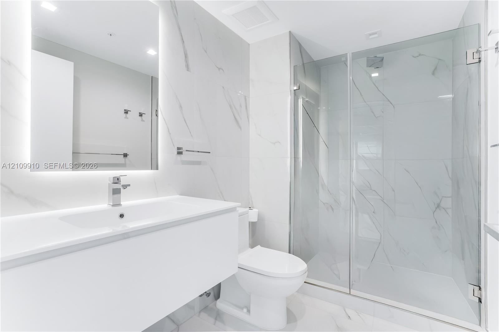 Bathroom featuring a floating vanity with a smooth surface, wall-to-wall mirror with integrated lighting, large-format stone-patterned wall tile, and a glass-enclosed shower