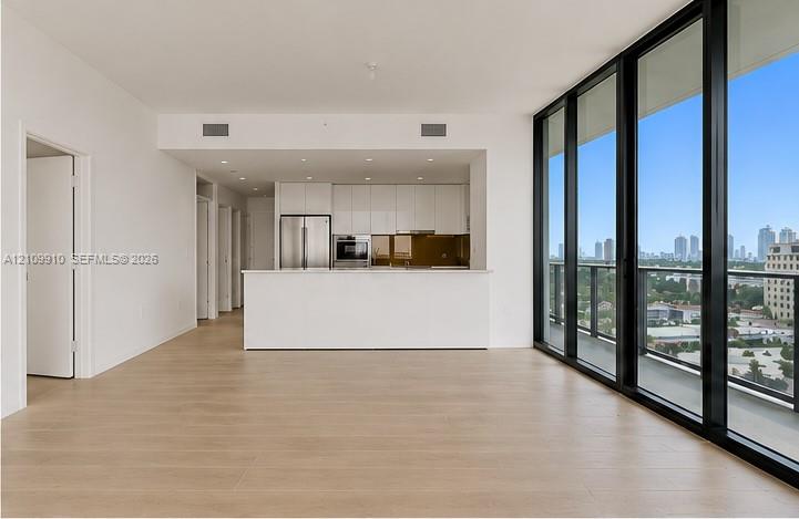 Open-concept interior featuring floor-to-ceiling glass panel walls, wood-finish flooring, white flat-panel cabinetry, stainless steel appliances, and a built-in kitchen peninsula