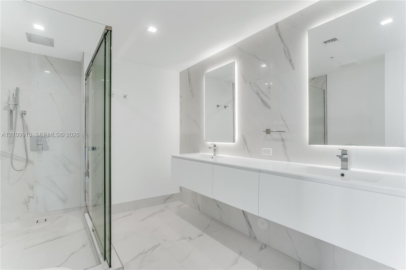 Floating double vanity with dual rectangular sinks, floor-to-ceiling stone-finish tile wall, glass-enclosed walk-in shower with rainfall fixture, and recessed LED-backlit mirrors