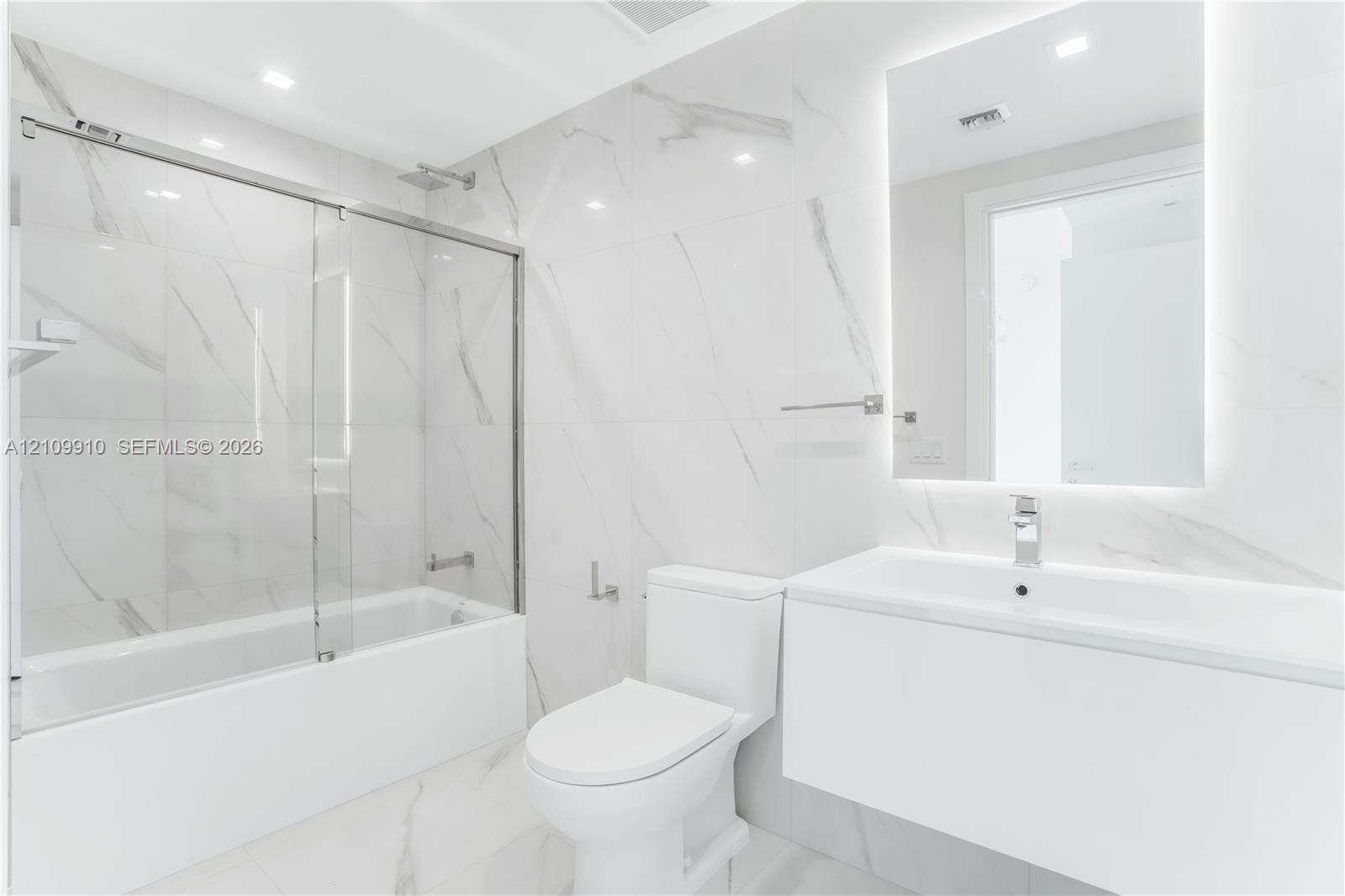 Bathroom featuring large-format porcelain tile, wall-mounted floating vanity, glass tub enclosure, and backlit vanity mirror