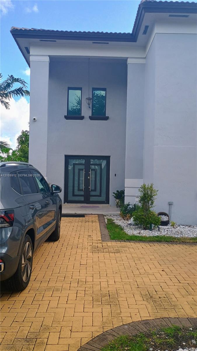 Two-story exterior featuring a light gray facade, paver driveway, double front entry doors with geometric glass inlays, and custom exterior lighting