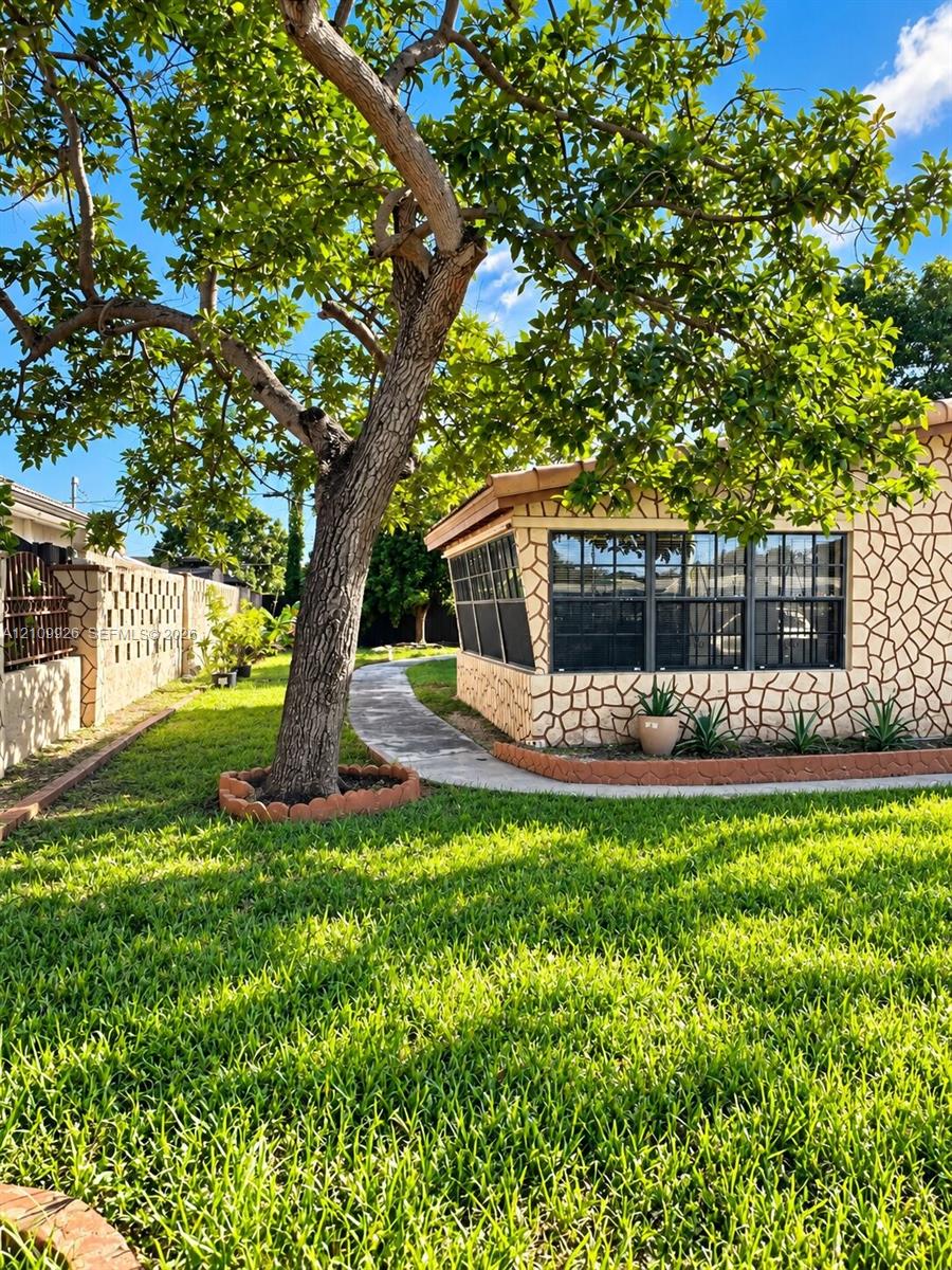 Spacious backyard featuring a manicured lawn, stone-patterned exterior masonry, mature canopy tree, concrete pathway, and concrete block perimeter wall