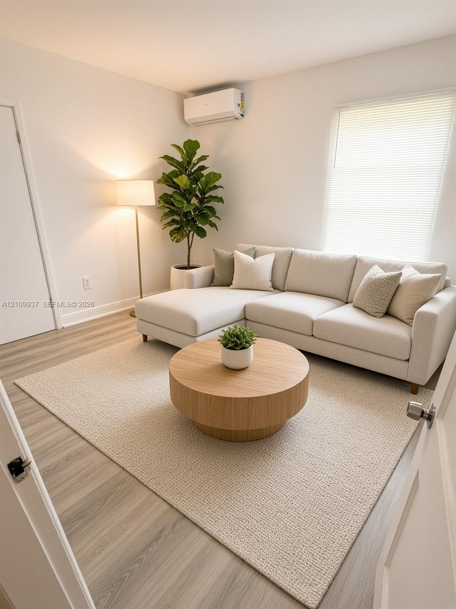 Neutral-toned interior featuring wood-finish flooring, a wall-mounted ductless climate control unit, and a window with horizontal blinds