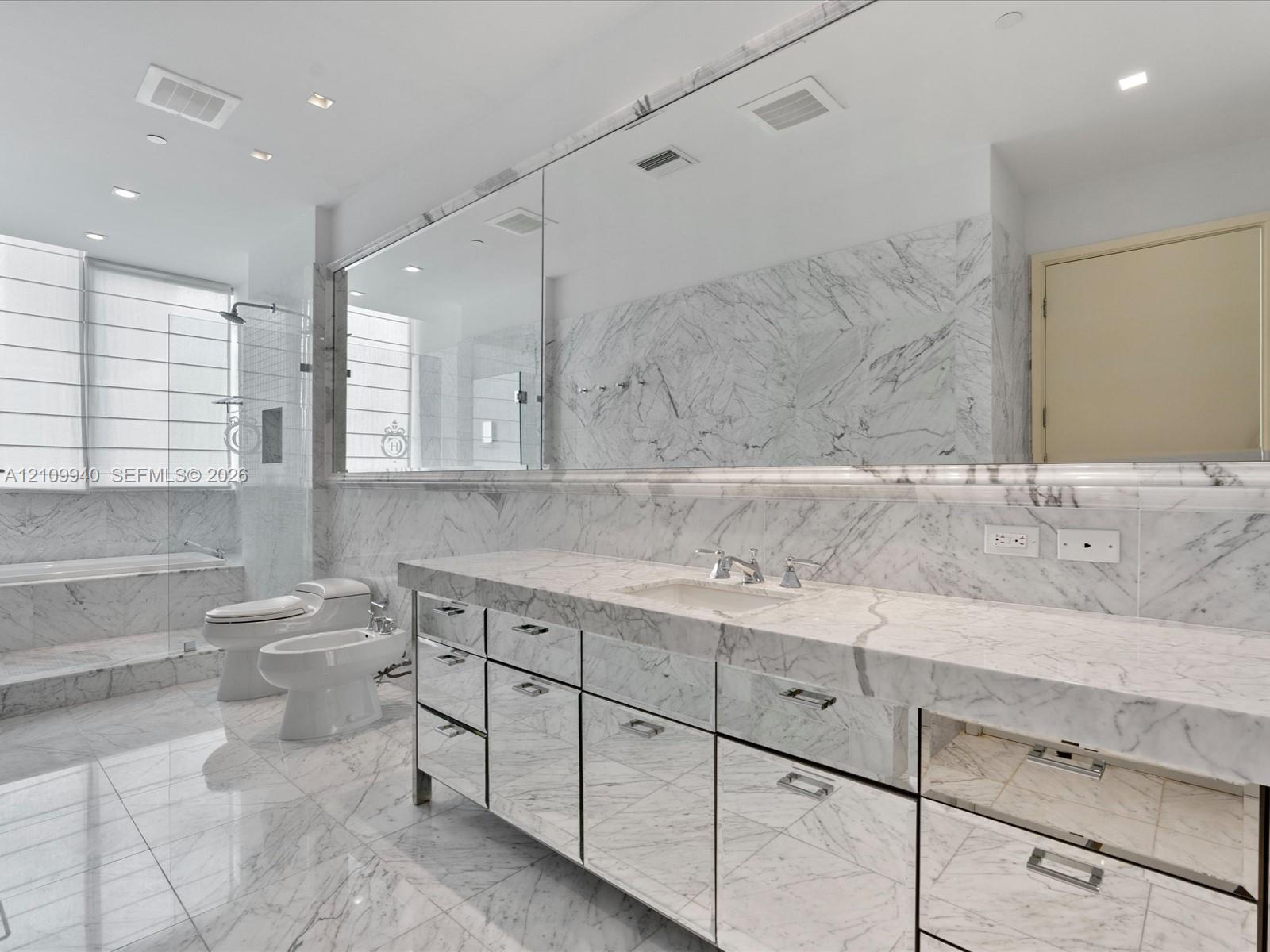 Full-wall vanity mirror, custom cabinetry with mirrored drawer fronts, natural stone countertops and wall cladding, recessed lighting, and a separate glass-enclosed shower and soaking tub