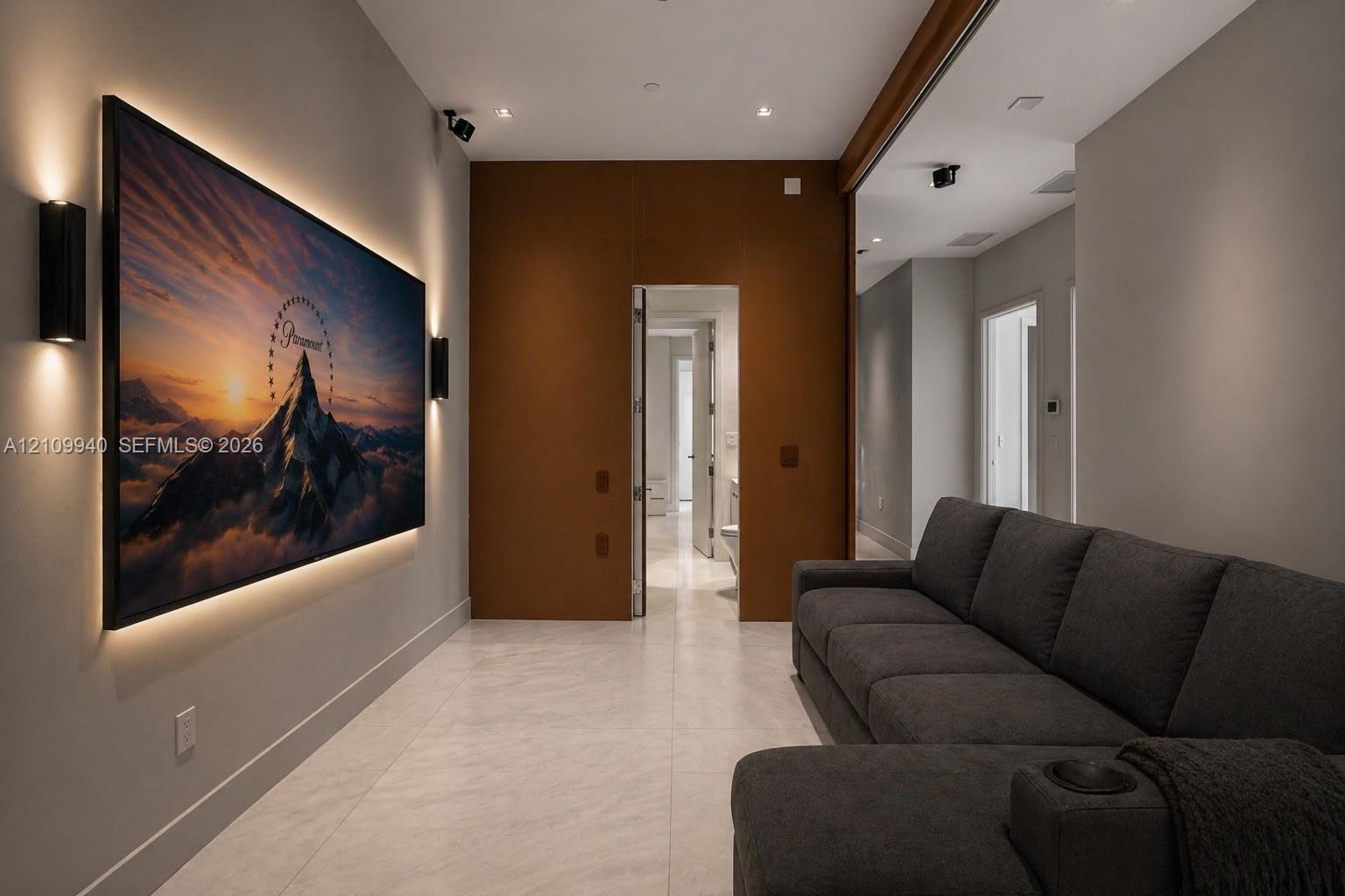 Media room featuring large-format wall-mounted screen with integrated LED backlighting, matte black wall-mounted sconces, recessed ceiling lighting, and smooth stone flooring