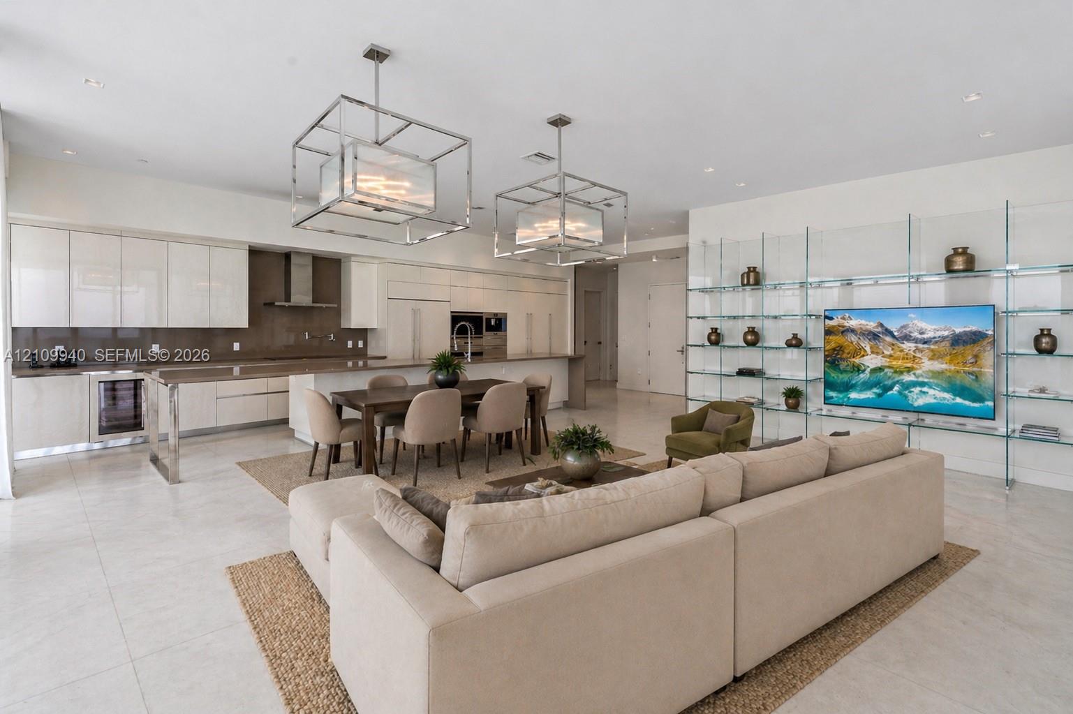 Open-concept area featuring high-gloss cabinetry, stainless steel built-in appliances, a large center island, dual geometric metal pendant lights, and floor-to-ceiling glass wall shelving
