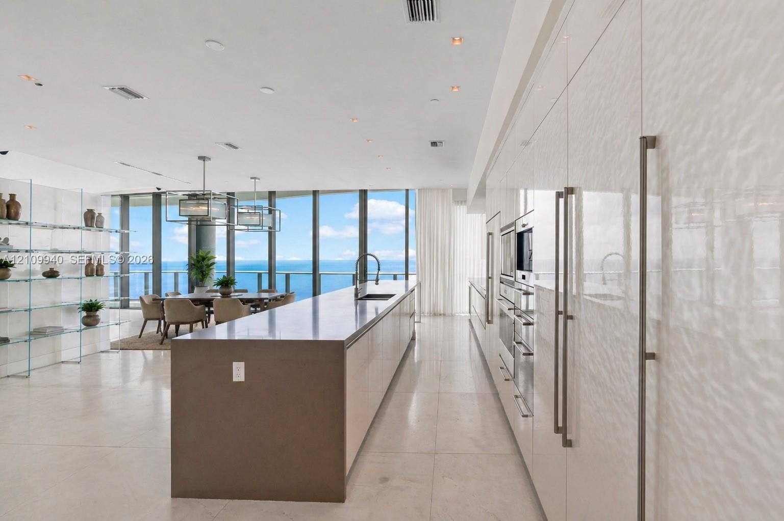 Modern kitchen featuring high-gloss cabinetry, stainless steel built-in appliances, a solid-surface center island with a waterfall edge, stone flooring, and floor-to-ceiling windows
