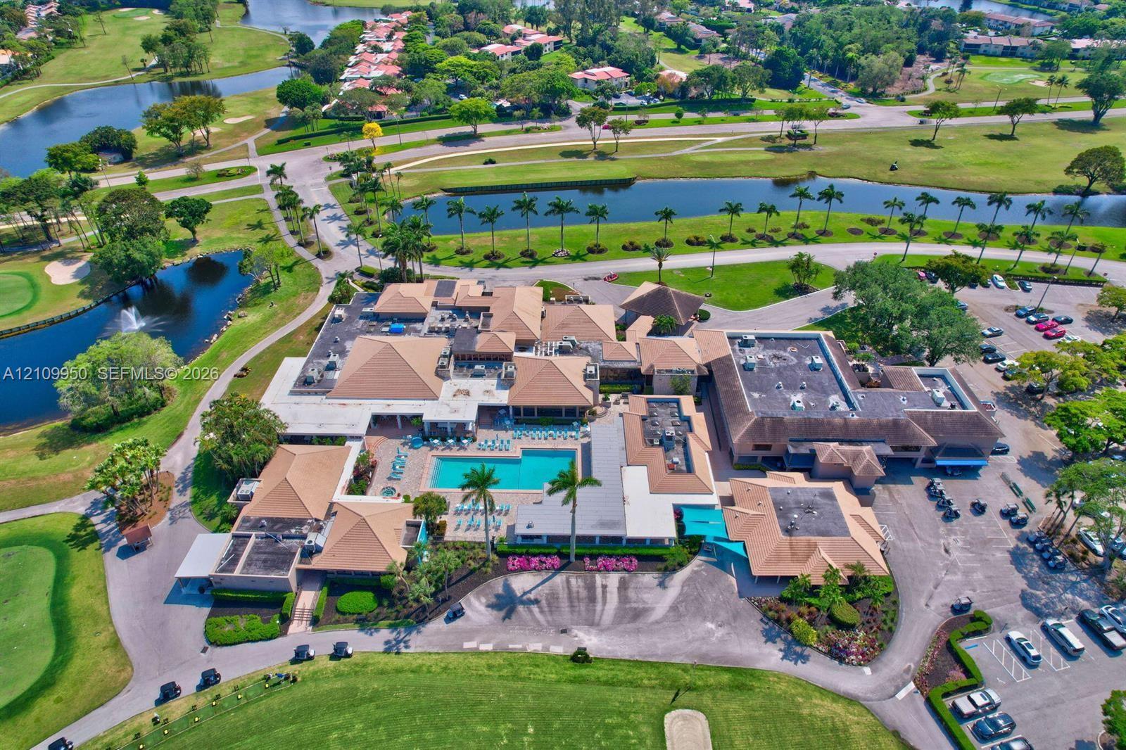 Aerial perspective of a multi-structure facility featuring a central swimming pool, paved surface parking, mature landscaping, and surrounding water features