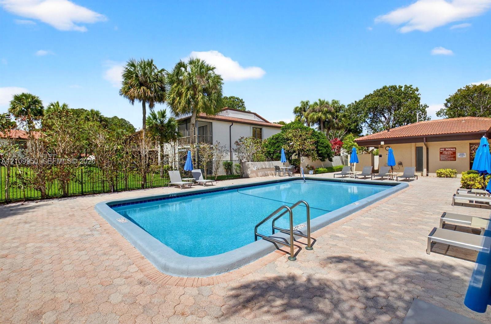 Rectangular swimming pool with metal handrails, surrounded by paver patio hardscaping, mature palm trees, and perimeter fencing