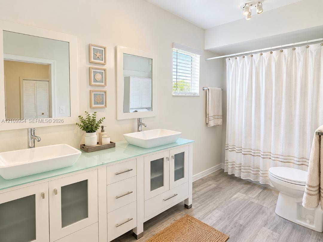 Bathroom featuring a dual-sink vanity with white cabinetry, frosted glass cabinet inserts, a light-toned countertop, rectangular vessel sinks, and wood-finish flooring Bathroom featuring a dual-sink vanity with white cabinetry, frosted glass cabinet inserts, a light-toned countertop, rectangular vessel sinks, and wood-finish flooring