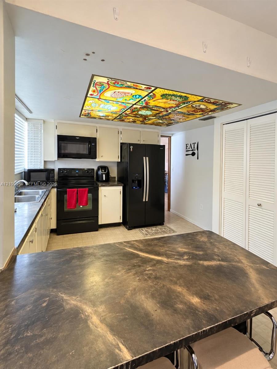 Kitchen featuring a decorative stained-glass ceiling fixture, dark-tone laminate countertops, solid-color cabinetry, black appliances, and louvered double-door storage Kitchen featuring a decorative stained-glass ceiling fixture, dark-tone laminate countertops, solid-color cabinetry, black appliances, and louvered double-door storage