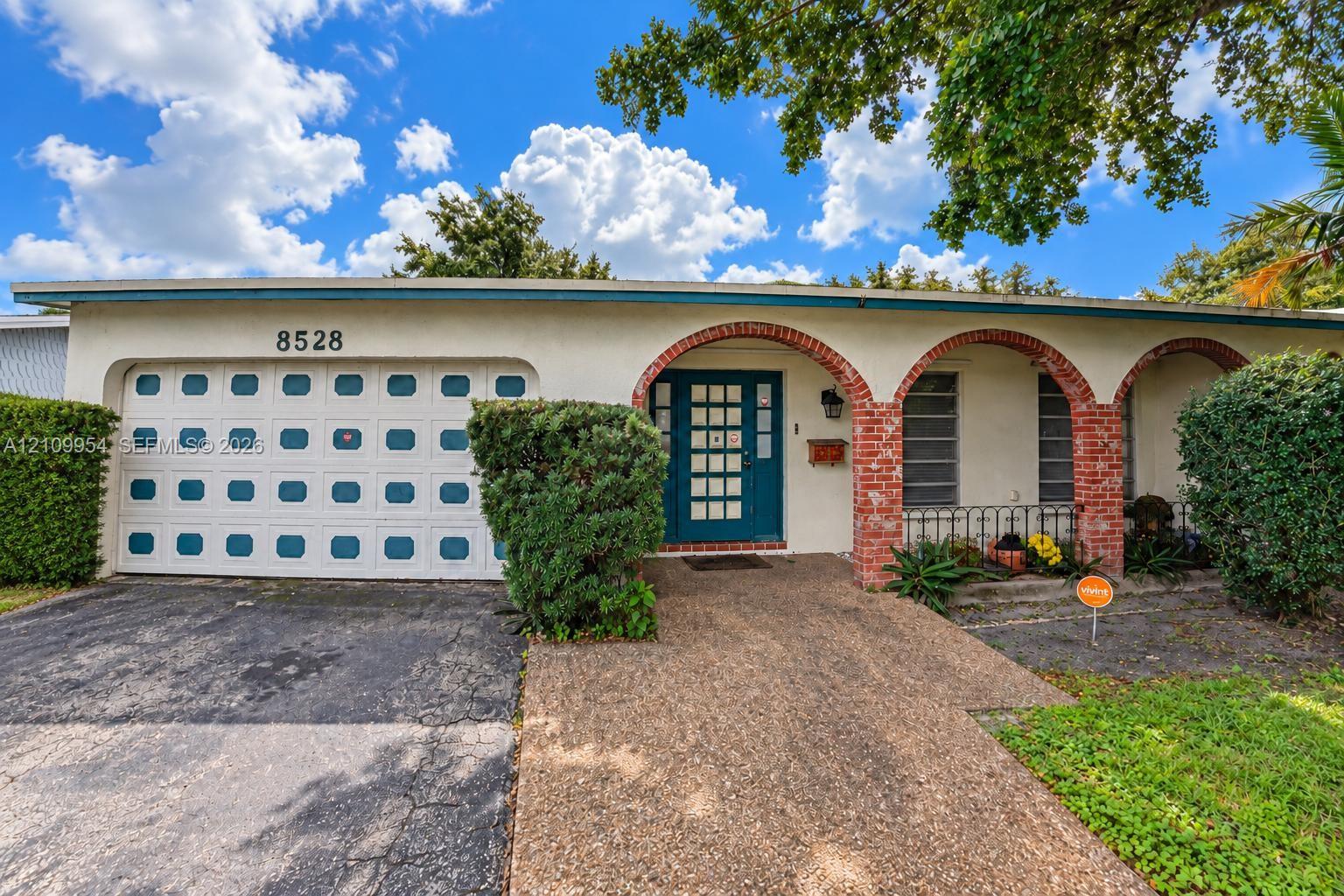 Single-story exterior featuring a stucco facade, red brick archway detail, aggregate concrete walkway, multi-pane front door, and a multi-panel garage door