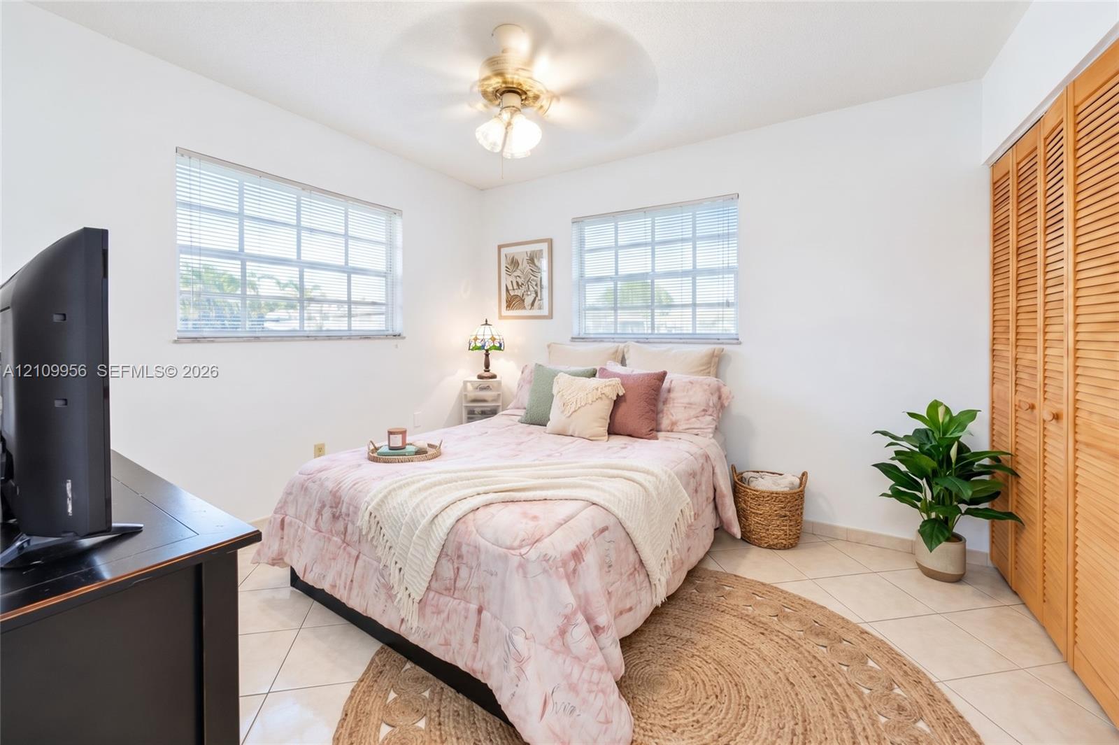 Interior space featuring light-toned tile flooring, dual windows with grid-pattern glass, a ceiling fan with integrated lighting, and full-height bi-fold louvered closet doors
