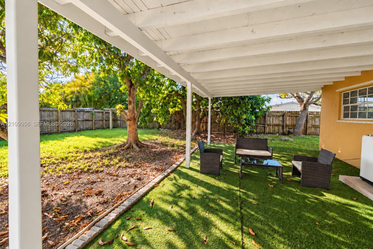 Covered patio featuring a white exposed-beam ceiling and wooden support posts, complemented by a fenced backyard with artificial turf, stone border edging, and mature trees