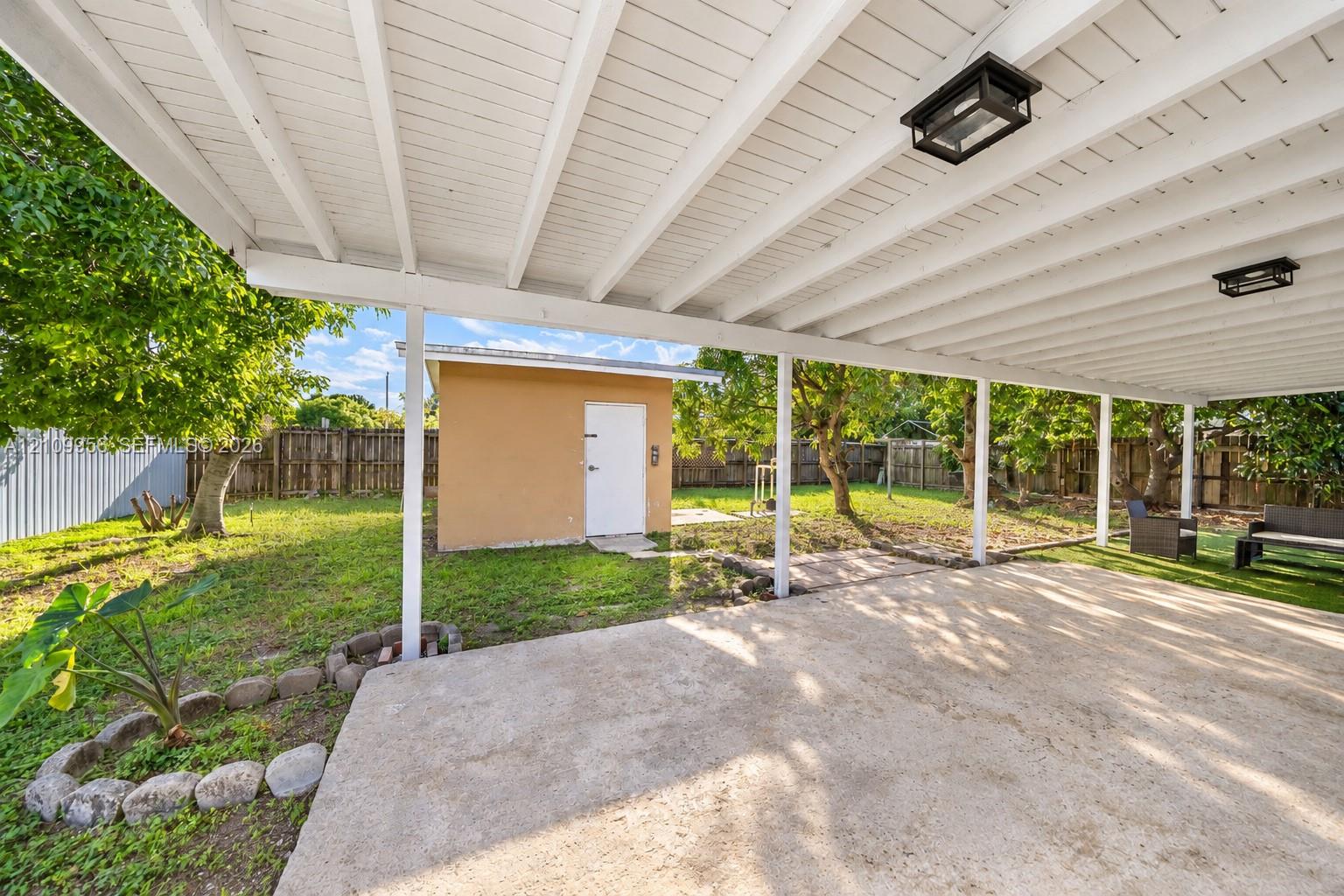 Covered concrete patio with overhead lighting, detached storage building, and fenced backyard with mature trees