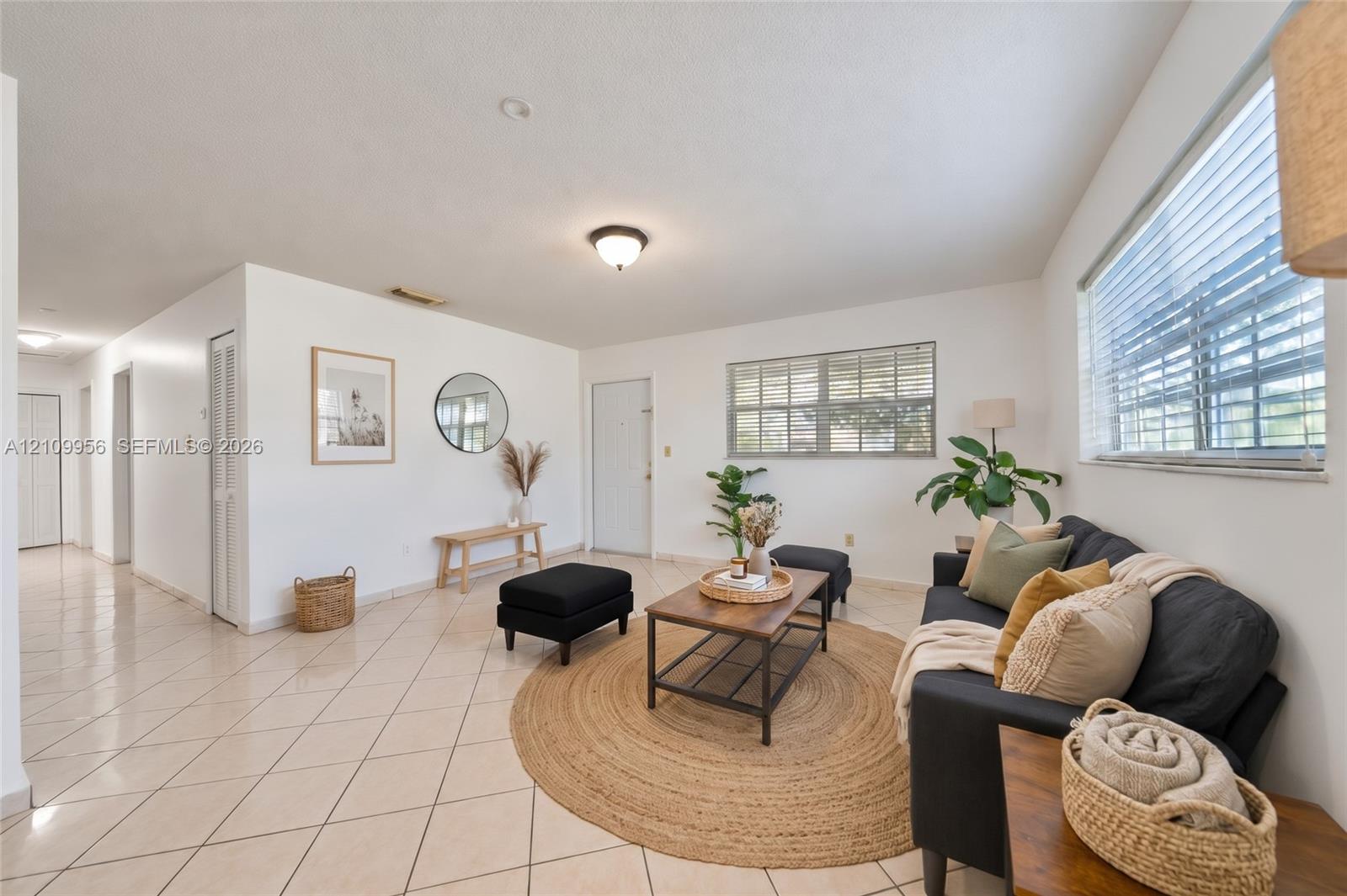 Open-concept interior featuring light-colored diagonal tile flooring, multi-pane windows with horizontal blinds, neutral wall paint, and overhead flush-mount lighting