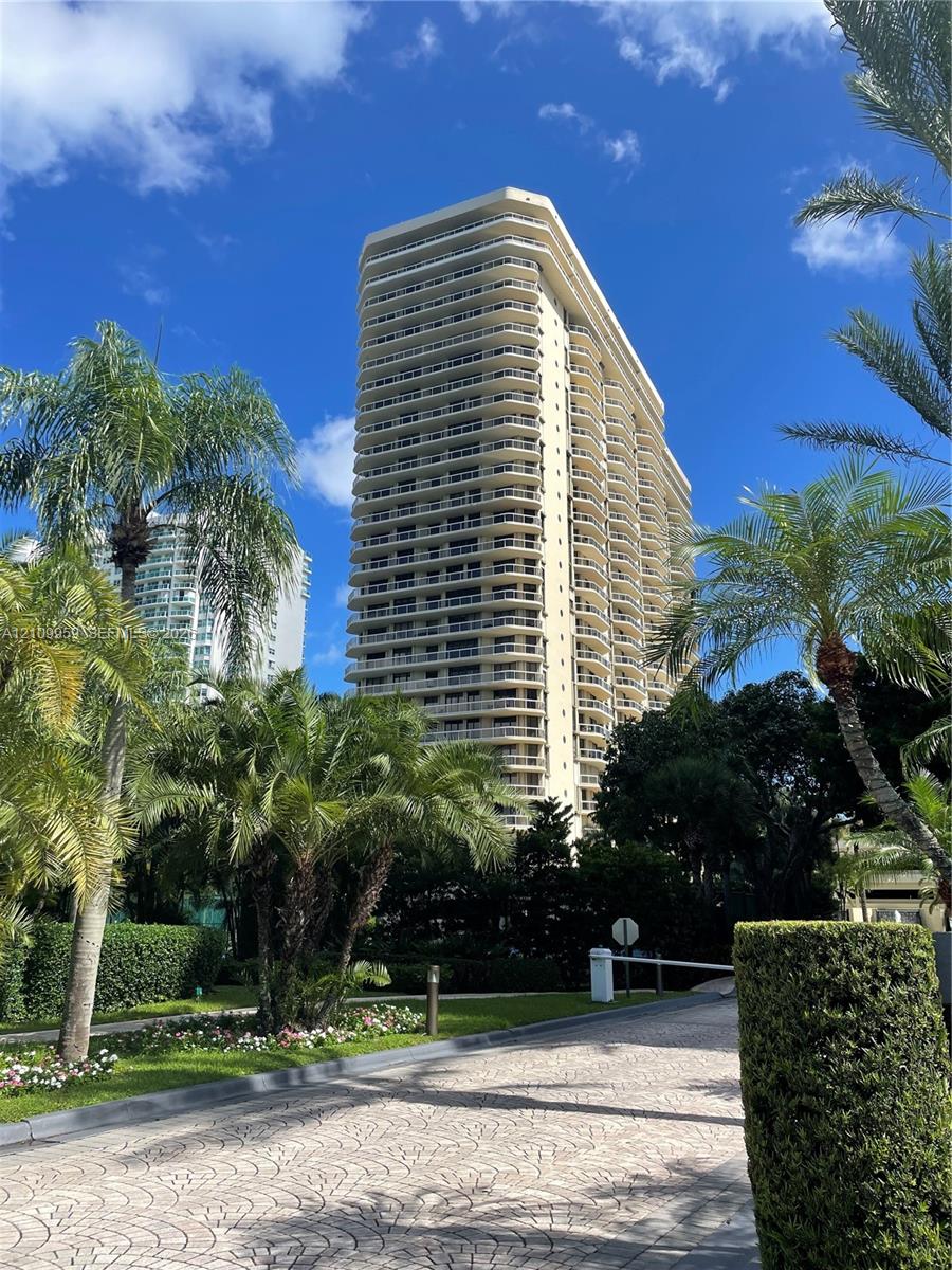 High-rise residential building featuring stacked private balconies, a paved drive, manicured tropical landscaping, and mature palm trees