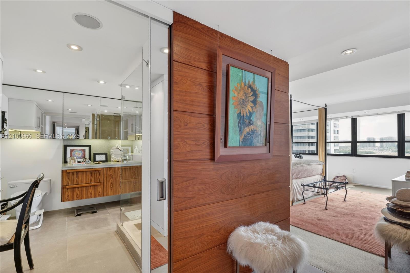 Floating wood-finish vanity with countertop, wood-panel accent wall with sliding door, recessed ceiling lighting, and large windows
