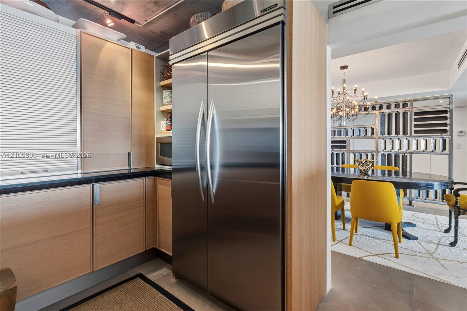 Kitchen featuring wood-veneer flat-panel cabinetry, professional-grade stainless steel refrigerator, black stone-composite countertops, and exposed concrete ceiling with track lighting
