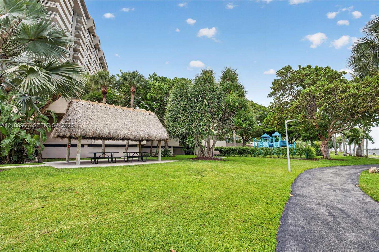 Landscaped grounds featuring a thatched-roof pavilion, paved walking paths, mature palm trees, and a play structure