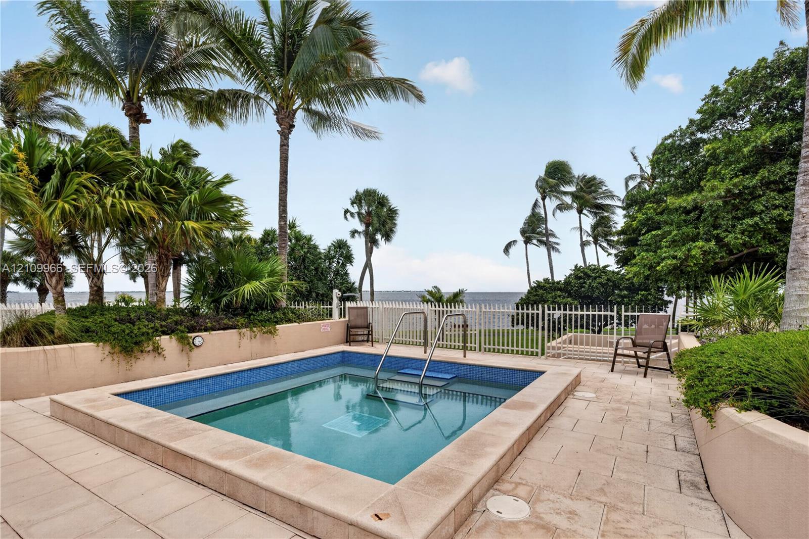 In-ground pool featuring blue tile inlay, stone-finish coping, metal handrails, and a surrounding light-toned stone deck with mature palm trees