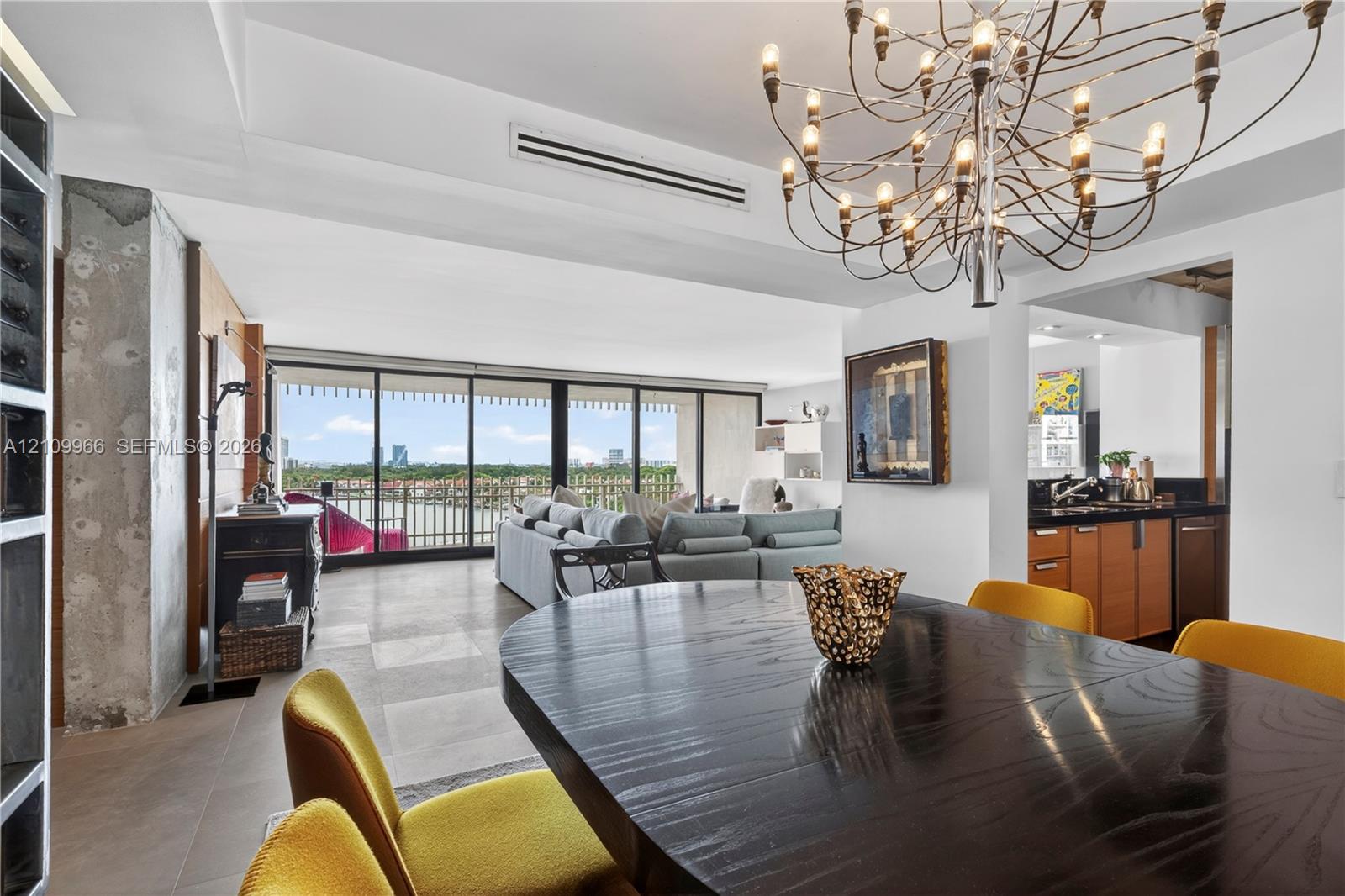 Open-concept interior featuring large-format tile flooring, exposed concrete structural column, floor-to-ceiling glass sliding doors, ornate metal chandelier, and built-in cabinetry