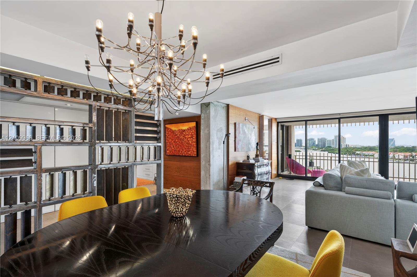 Open-concept interior featuring a multi-arm modern chandelier, custom decorative metal partition wall, wood-paneled feature wall, floor-to-ceiling glass sliding doors, and light-toned tile flooring