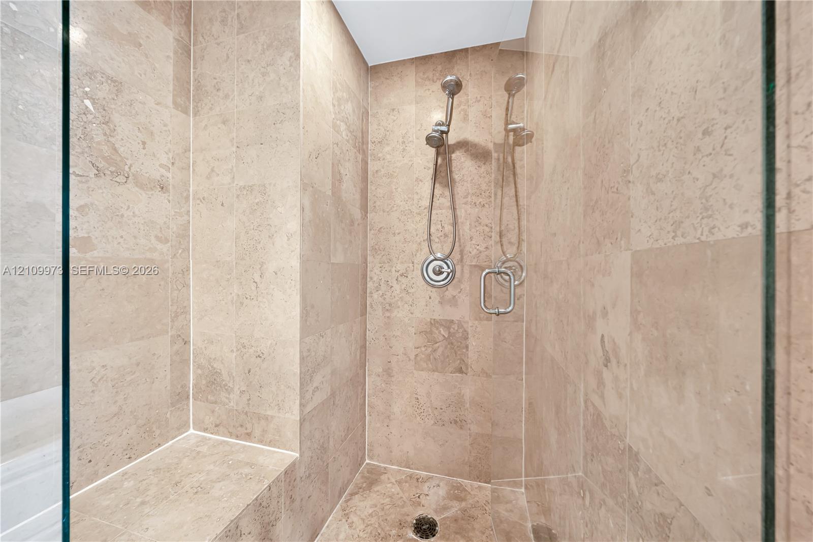 Walk-in shower featuring floor-to-ceiling stone tile, built-in stone bench, glass enclosure, and adjustable handheld shower fixture