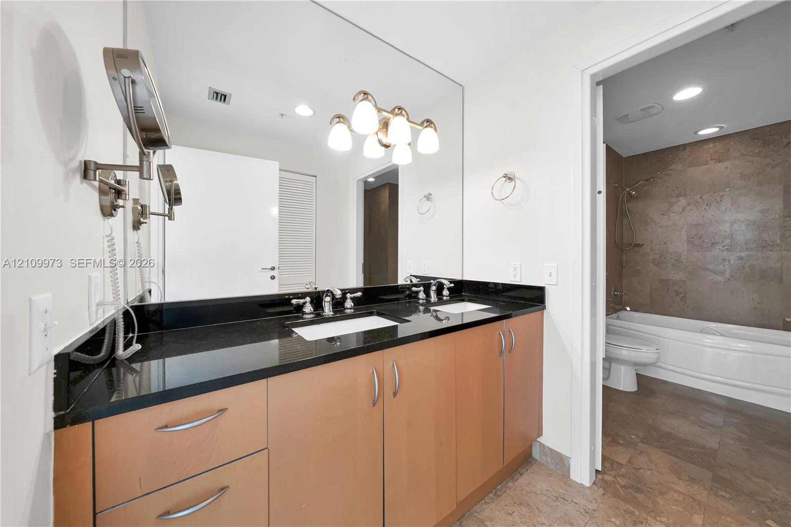 Dual-sink vanity featuring a dark stone countertop, light wood cabinetry, a large wall-to-wall mirror, and multi-light vanity lighting
