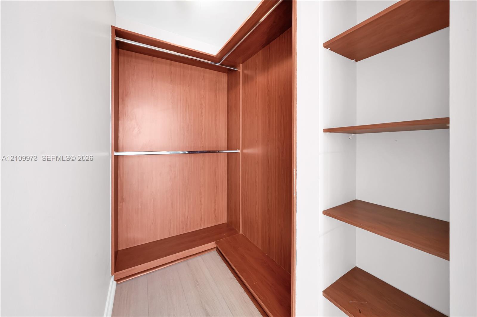 Built-in closet featuring wood-finish shelving, an L-shaped hanging rod system, and neutral wood-finish flooring