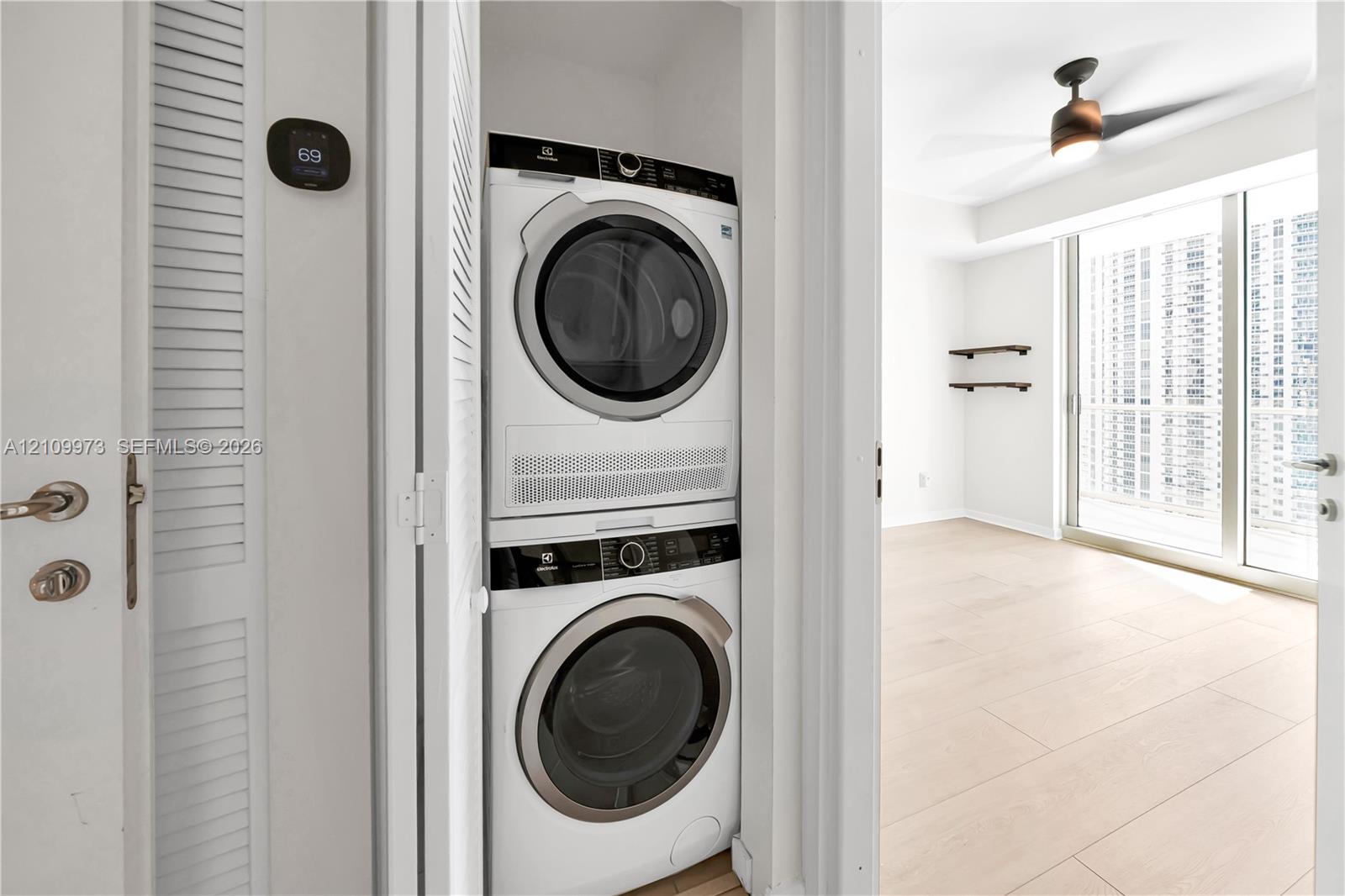 Stackable laundry unit featuring an Electrolux washer and dryer, digital wall-mounted thermostat, wood-finish flooring, ceiling fan with integrated lighting, and wall-mounted shelving