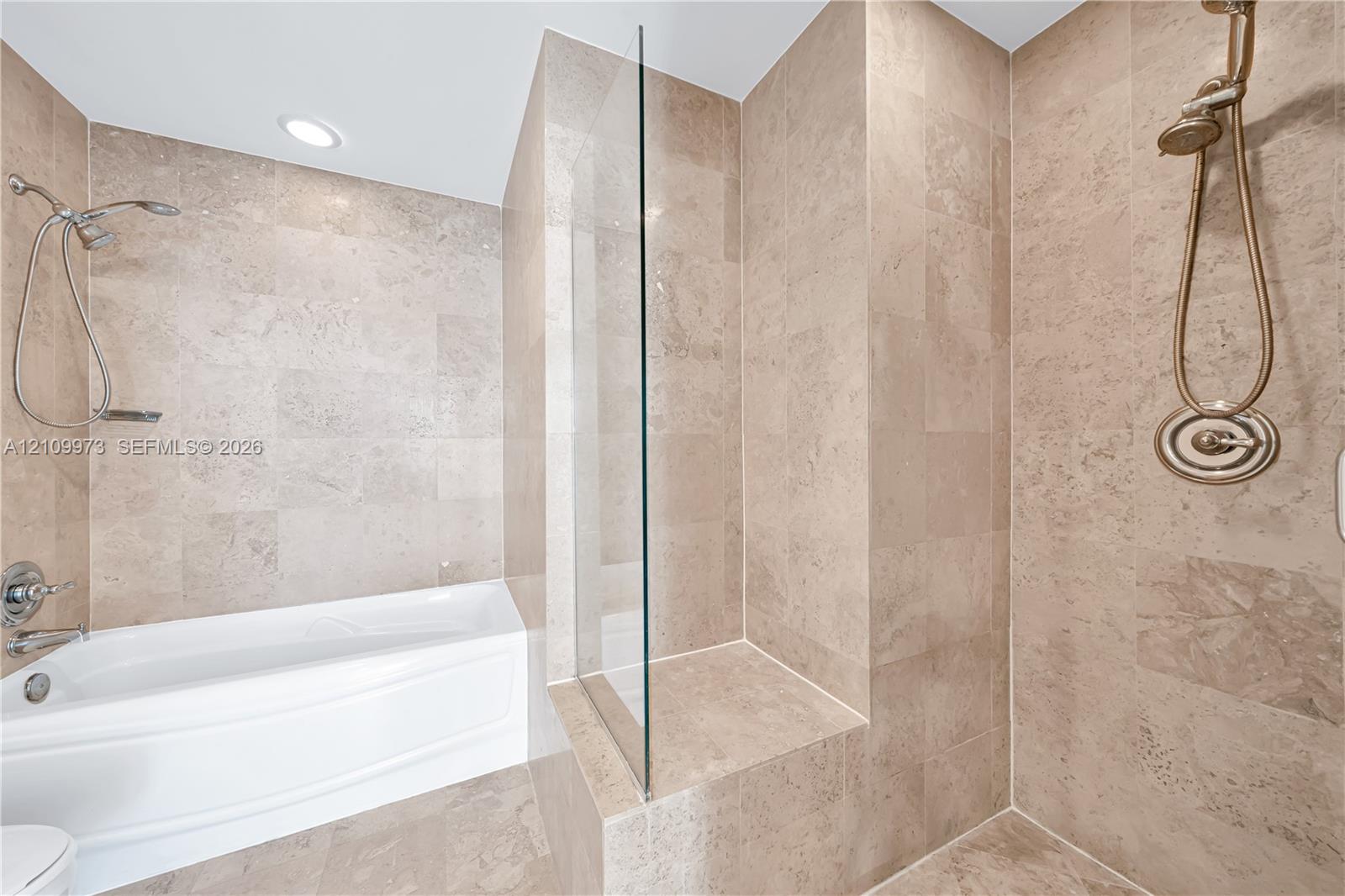 Soaking tub with a wall-mounted showerhead, separate glass-enclosed shower featuring a built-in tiled bench, and neutral stone-finish tile surround