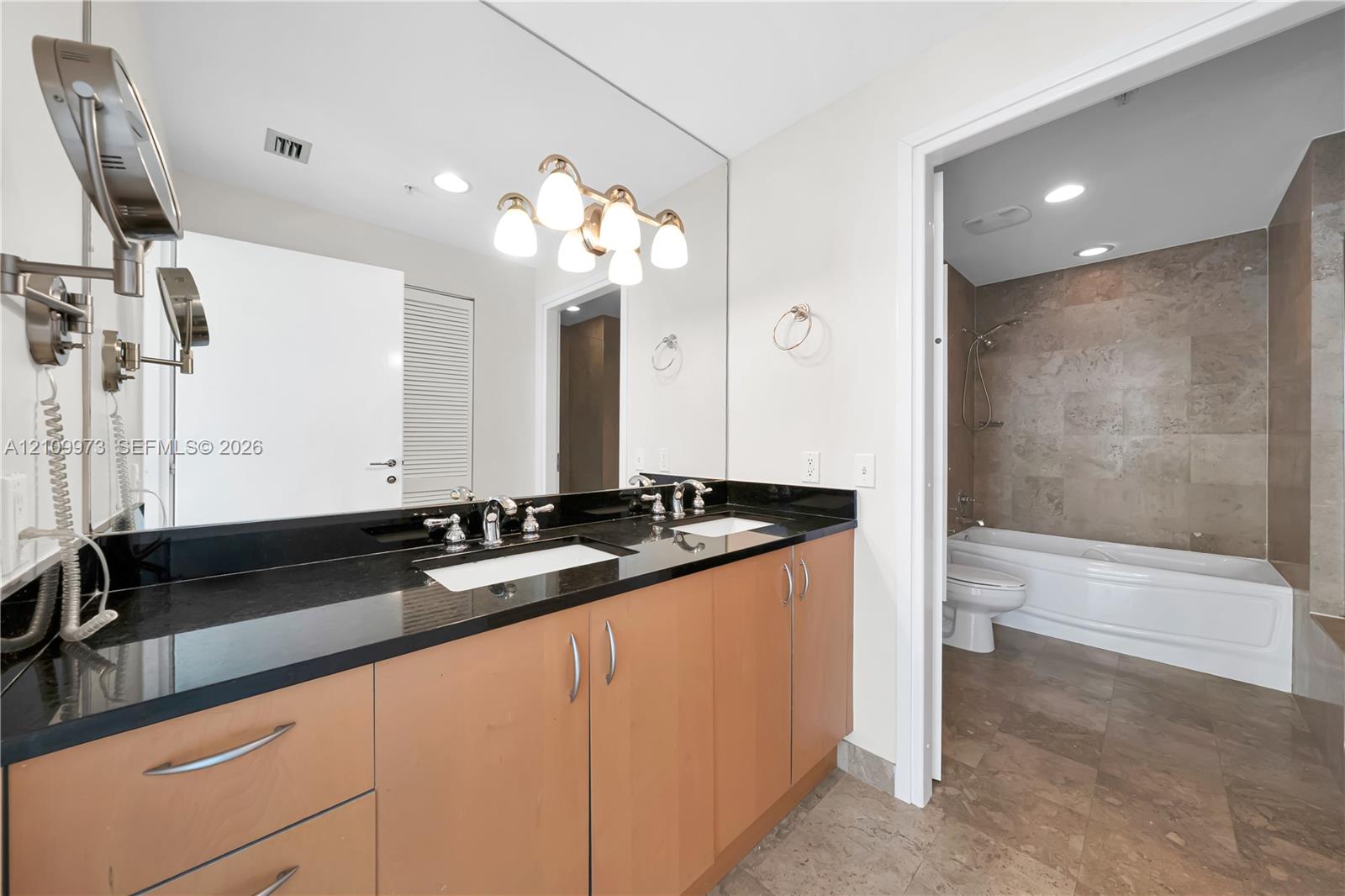 Dual-sink vanity with dark stone countertop, wood-finish cabinetry, full-width mirror, and multi-bulb vanity lighting, featuring a tile-surround bathtub