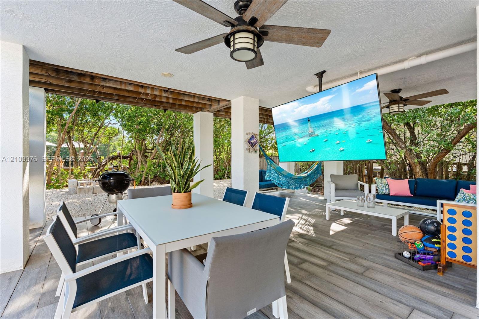 Covered outdoor living area featuring wood-finish tile flooring, ceiling fans with integrated lighting, and white structural columns