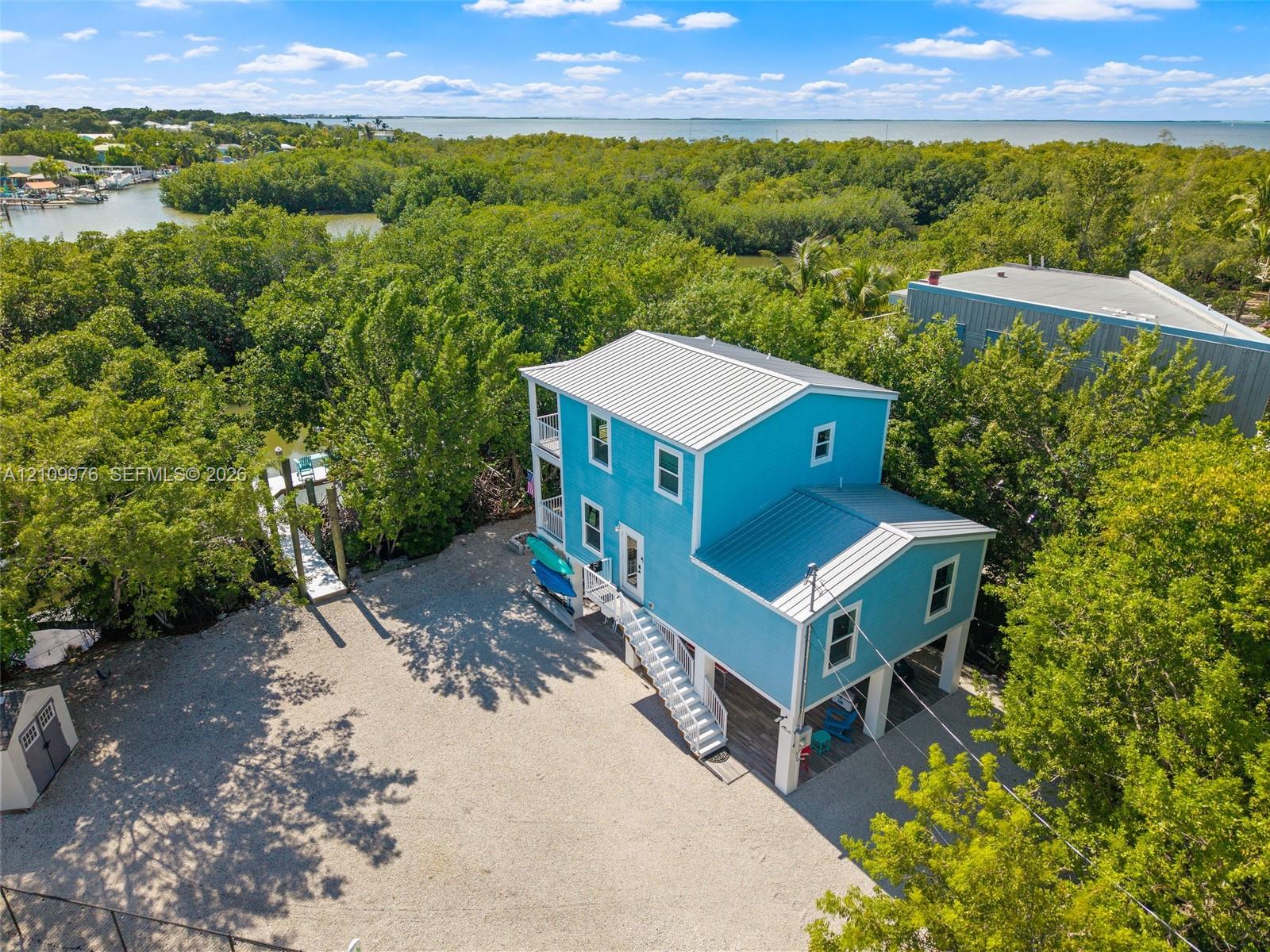 Two-story elevated coastal residence featuring a metal roof, wraparound exterior staircases, covered ground-level parking, gravel driveway, and a private dock