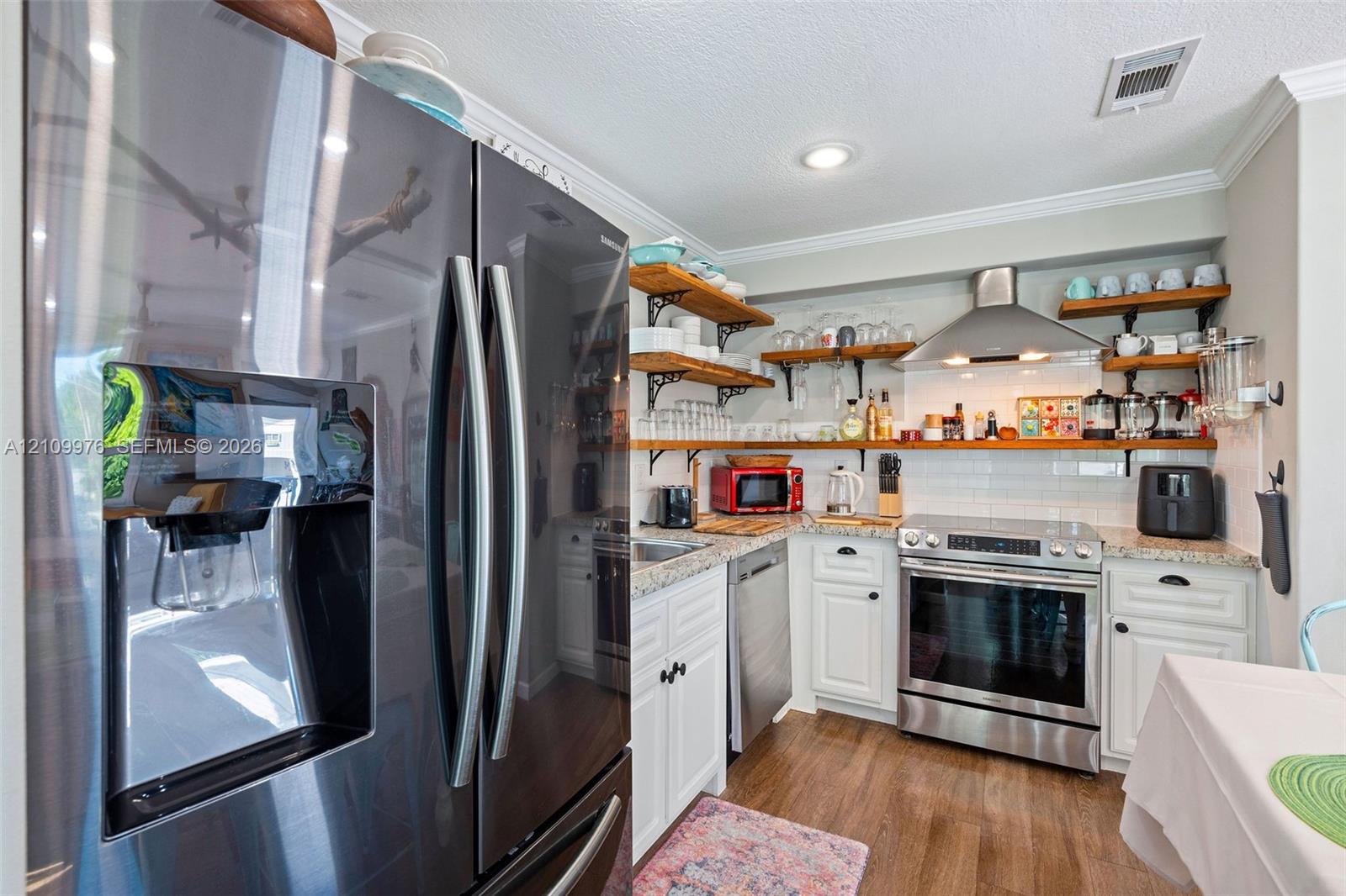 Kitchen featuring stainless steel appliances, stone-look countertops, white cabinetry with crown molding, natural wood-finish open shelving, and a white subway tile backsplash