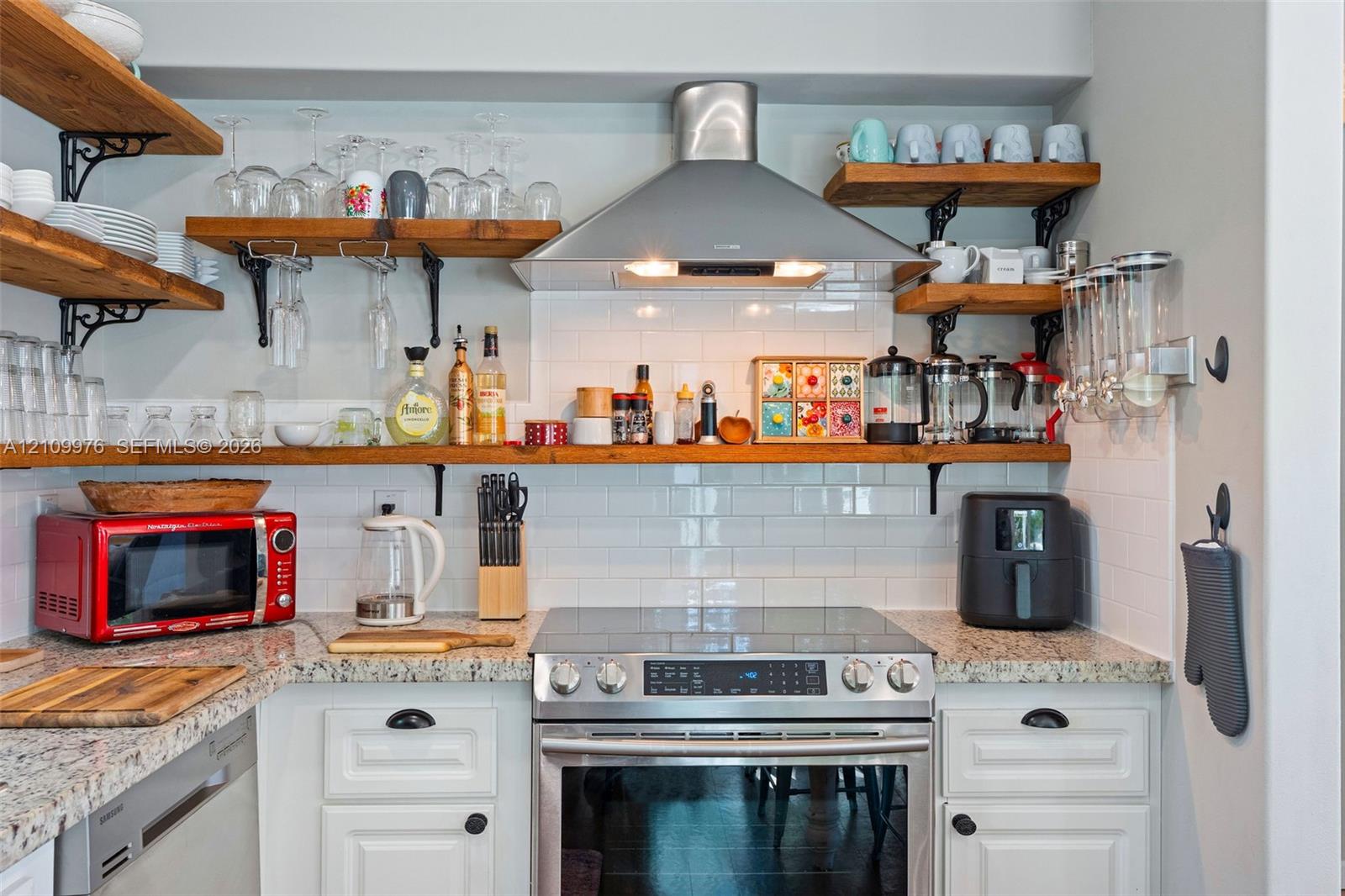 Kitchen area featuring white shaker-style cabinetry, natural stone countertops, white subway tile backsplash, stainless steel range with overhead vent hood, and open wood shelving