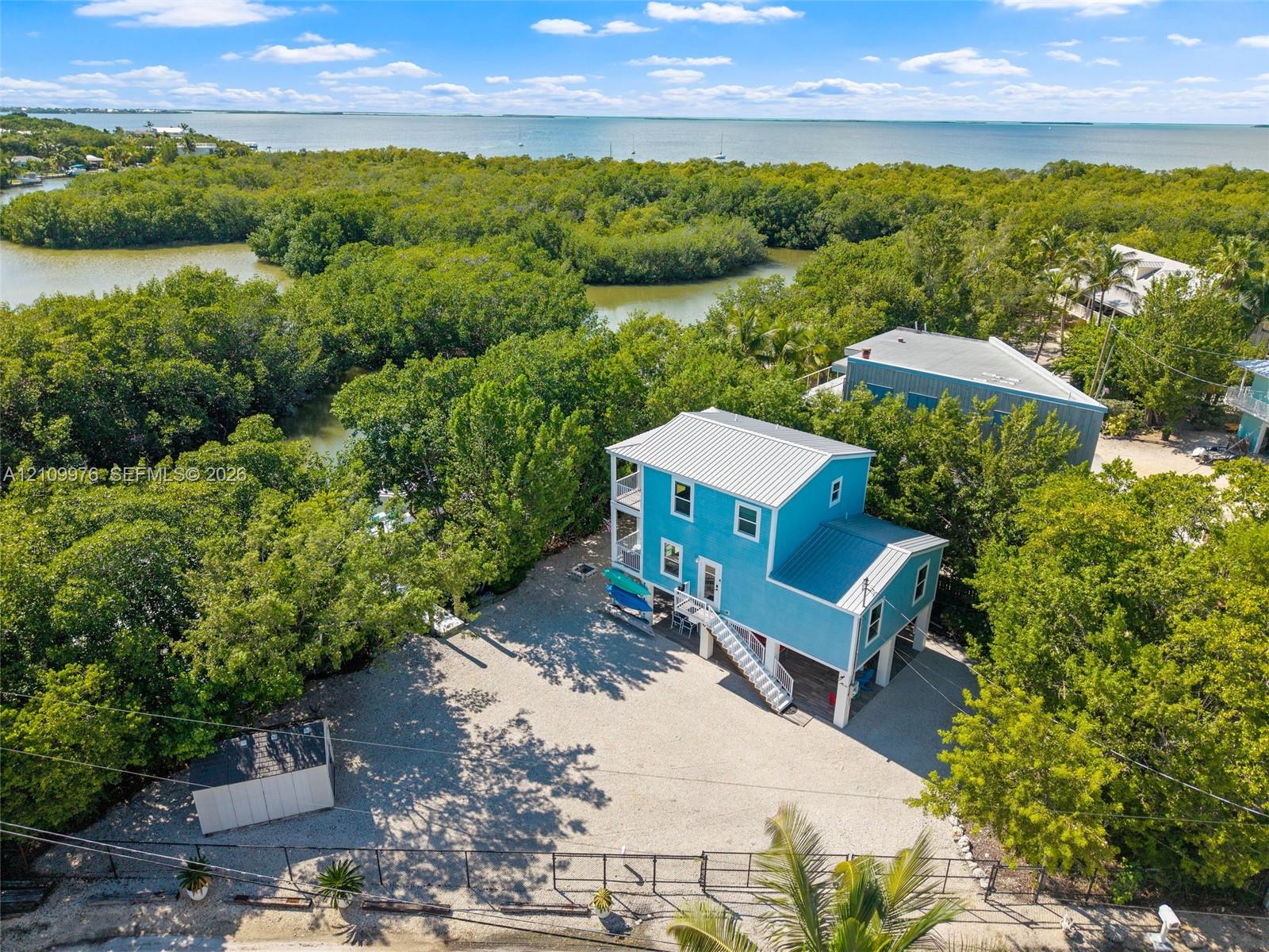 Elevated residence featuring a metal roof, exterior stairway, stone-surface driveway, and onsite storage structure, situated within proximity to coastal waterways and mangroves