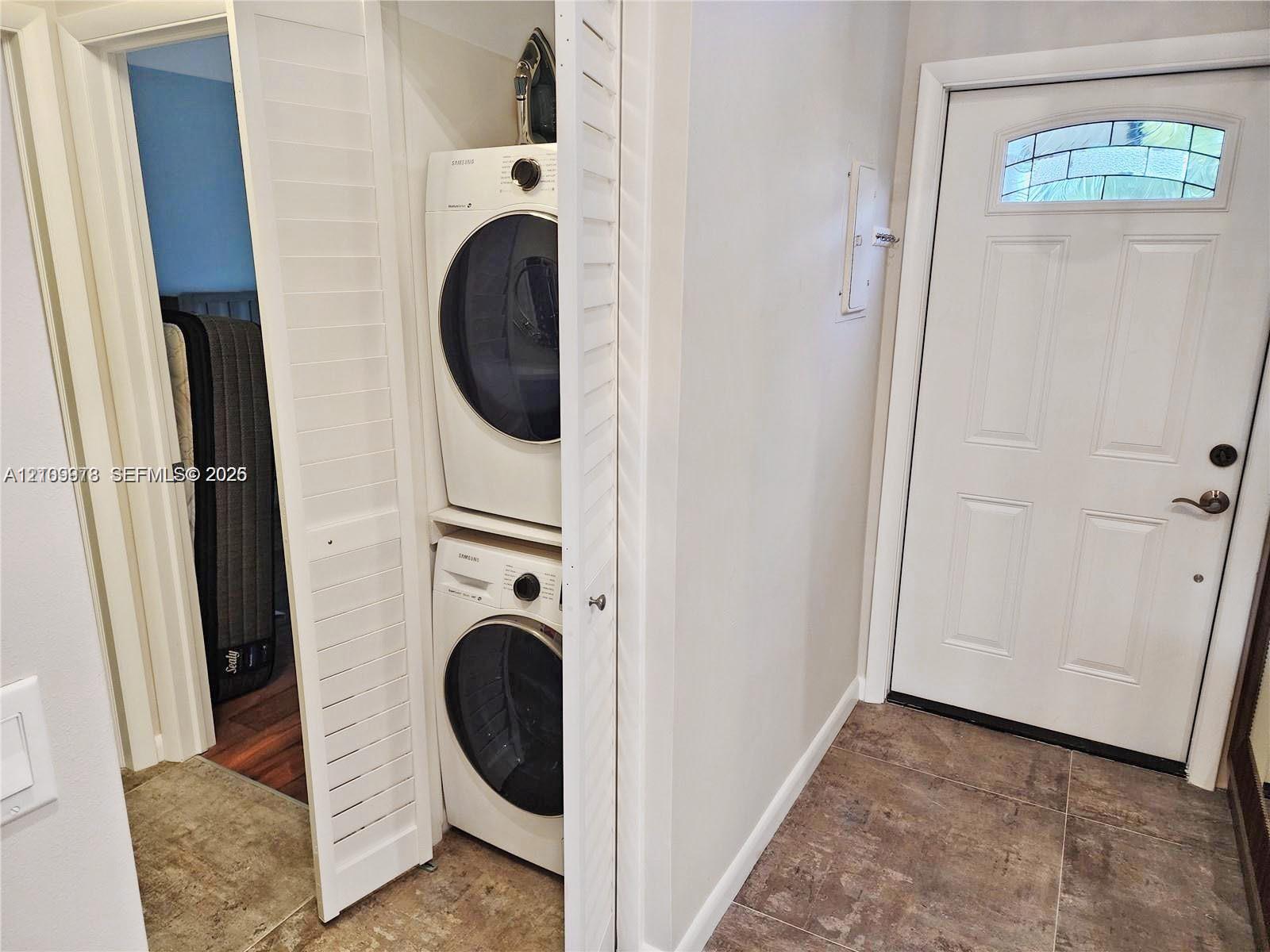 Laundry closet featuring stacked front-load washer and dryer, louvered bi-fold doors, and a exterior-facing door with an arched glass inset