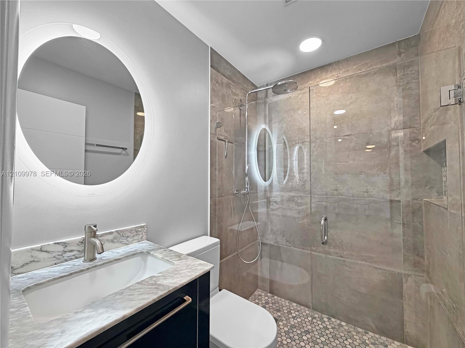 Modern bathroom featuring a circular LED-backlit mirror, vanity with stone countertop, glass-enclosed shower with large-format wall tiling, and mosaic floor tile