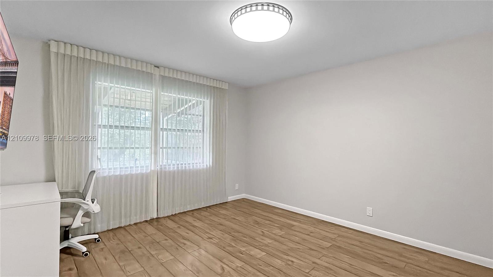 Neutral-toned interior featuring wood-finish flooring, a flush-mount circular ceiling fixture, and floor-to-ceiling pleated window treatments