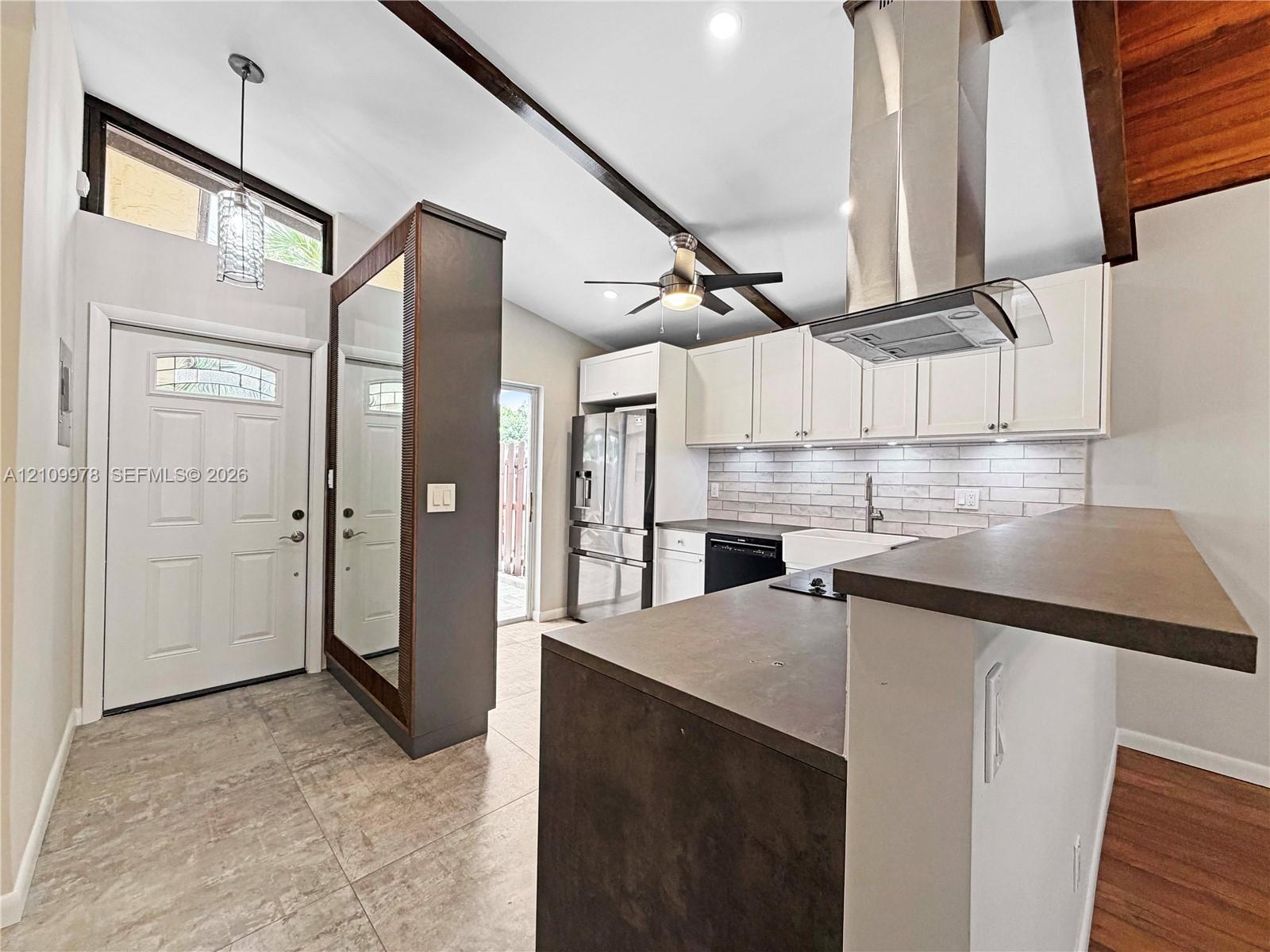 Vaulted ceilings with exposed structural beams, kitchen featuring white shaker-style cabinetry, matte finish countertop with breakfast bar, stainless steel chimney-style range hood, and tile flooring Vaulted ceilings with exposed structural beams, kitchen featuring white shaker-style cabinetry, matte finish countertop with breakfast bar, stainless steel chimney-style range hood, and tile flooring