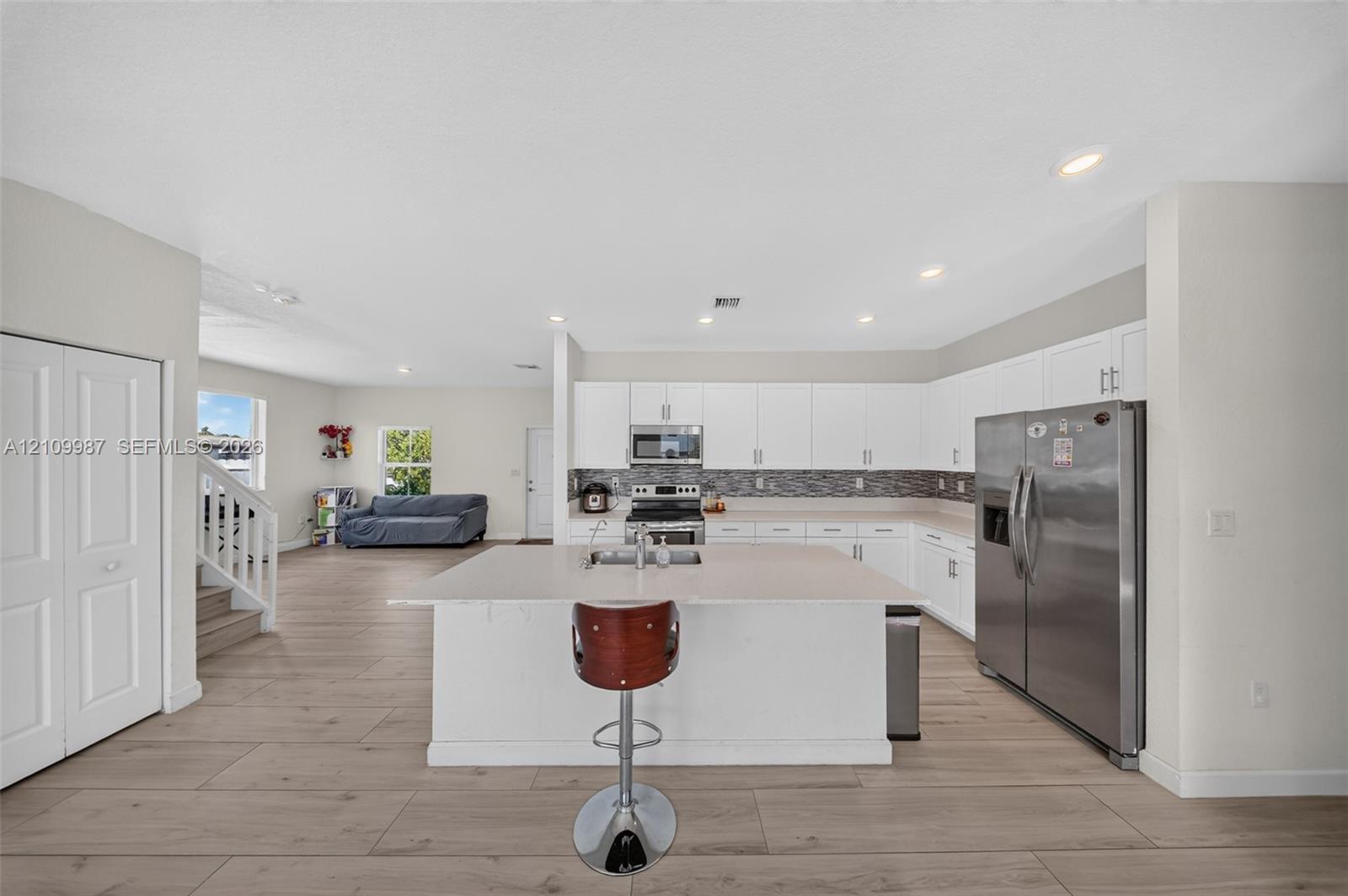 Kitchen featuring white shaker-style cabinetry, quartz countertops, a central island with an integrated sink, stainless steel appliances, and porcelain flooring