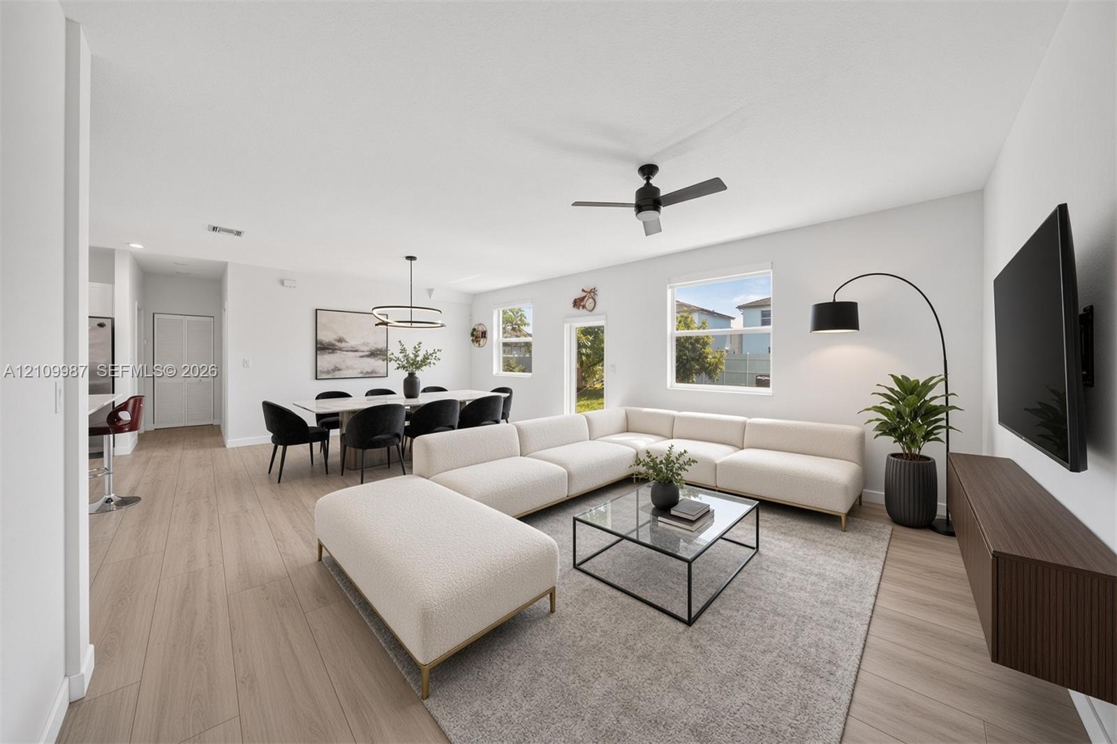 Staged Open-concept interior featuring porcelain flooring, ceiling fan with integrated lighting, dual-tiered modern chandelier, and a wall-mounted floating cabinetry unit