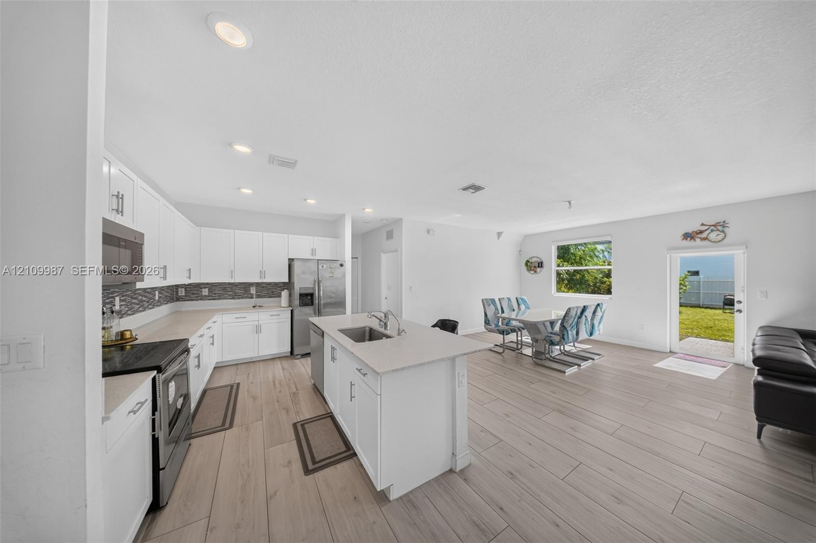 Kitchen featuring white cabinetry, center island with stainless steel sink, quartz countertops, tile backsplash, and porcelain floor.