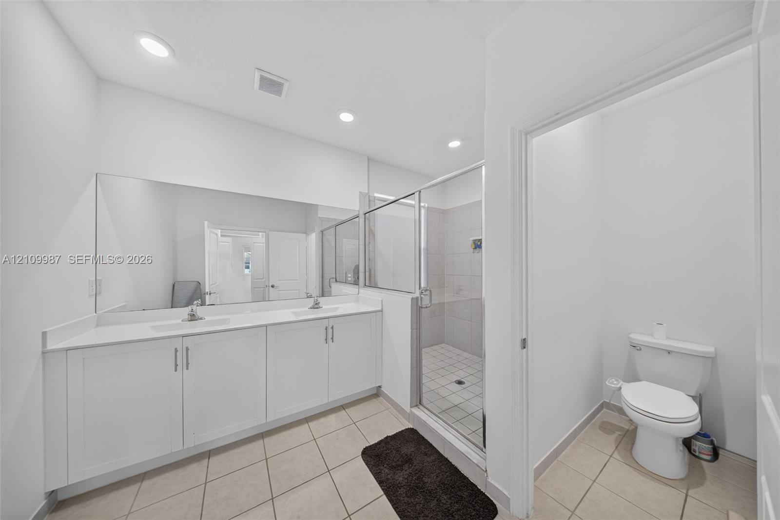 Dual-sink vanity with white cabinetry, expansive wall-to-wall mirror, walk-in shower with glass enclosure, and tile flooring