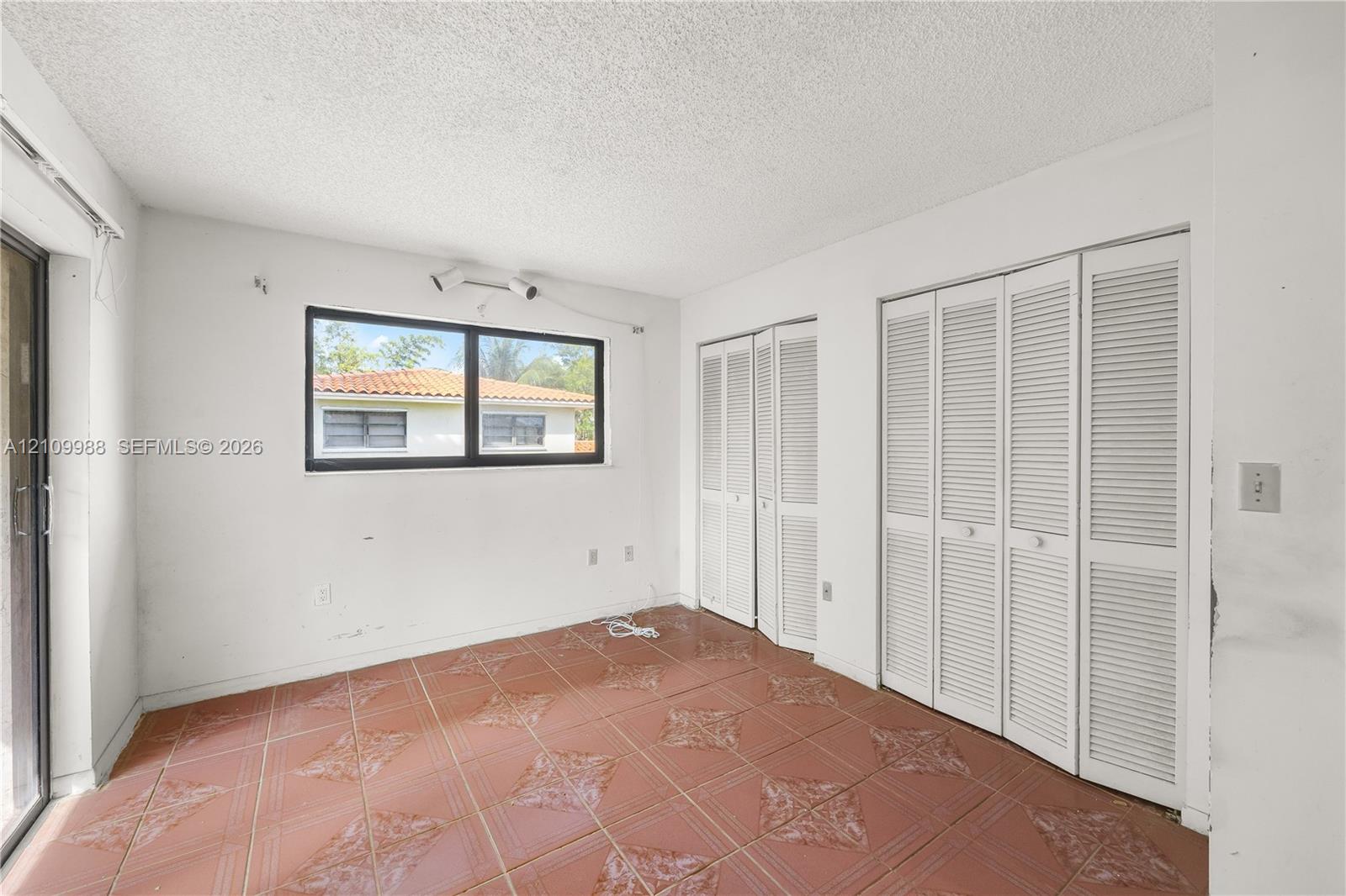 Room featuring terracotta-style tile flooring, dual sets of white louvered-door closets, and a large window with black frames